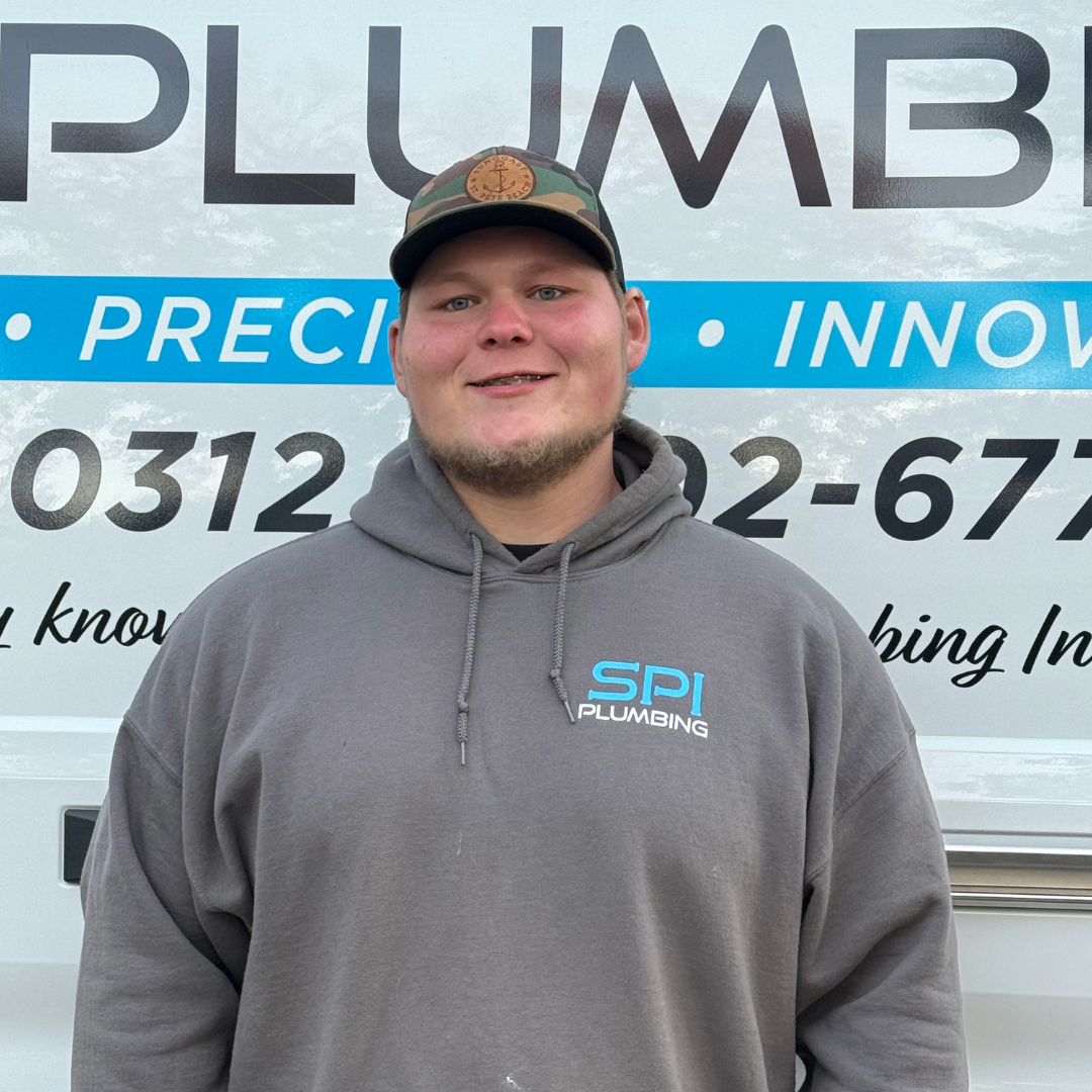 A young man wearing a gray hoodie with 'SPI Plumbing' logo standing in front of a company's service truck with signs and contact information.