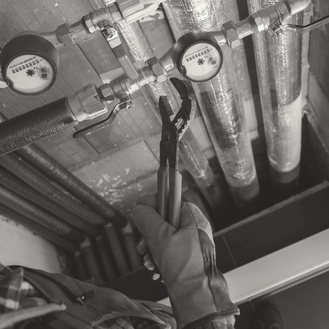 Close-up of a worker's gloved hand holding pliers near a water meter and plumbing pipes in a ceiling.