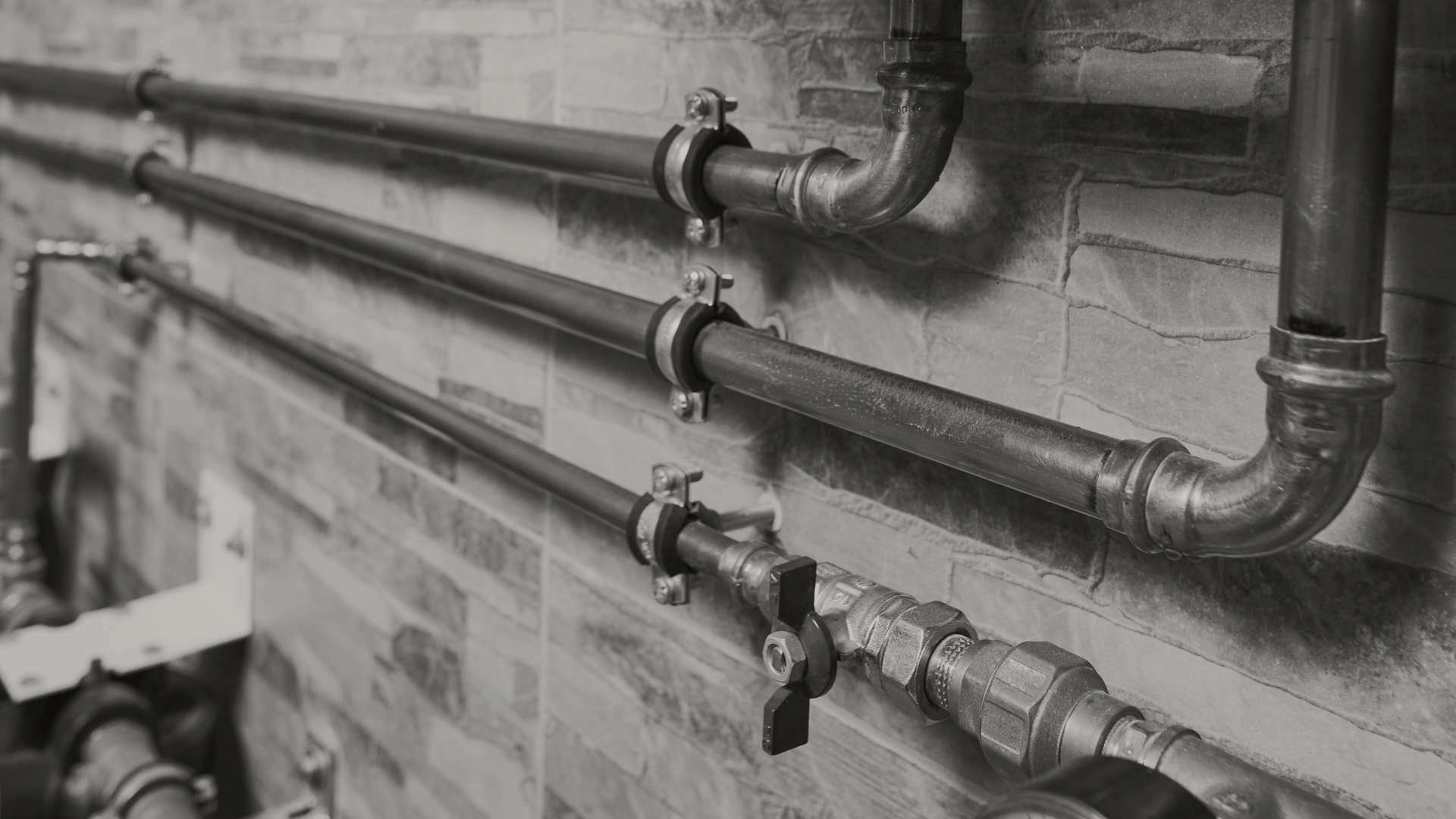 Black and white image of industrial metal pipes mounted on a brick wall, secured with circular clamps and a valve for regulation.