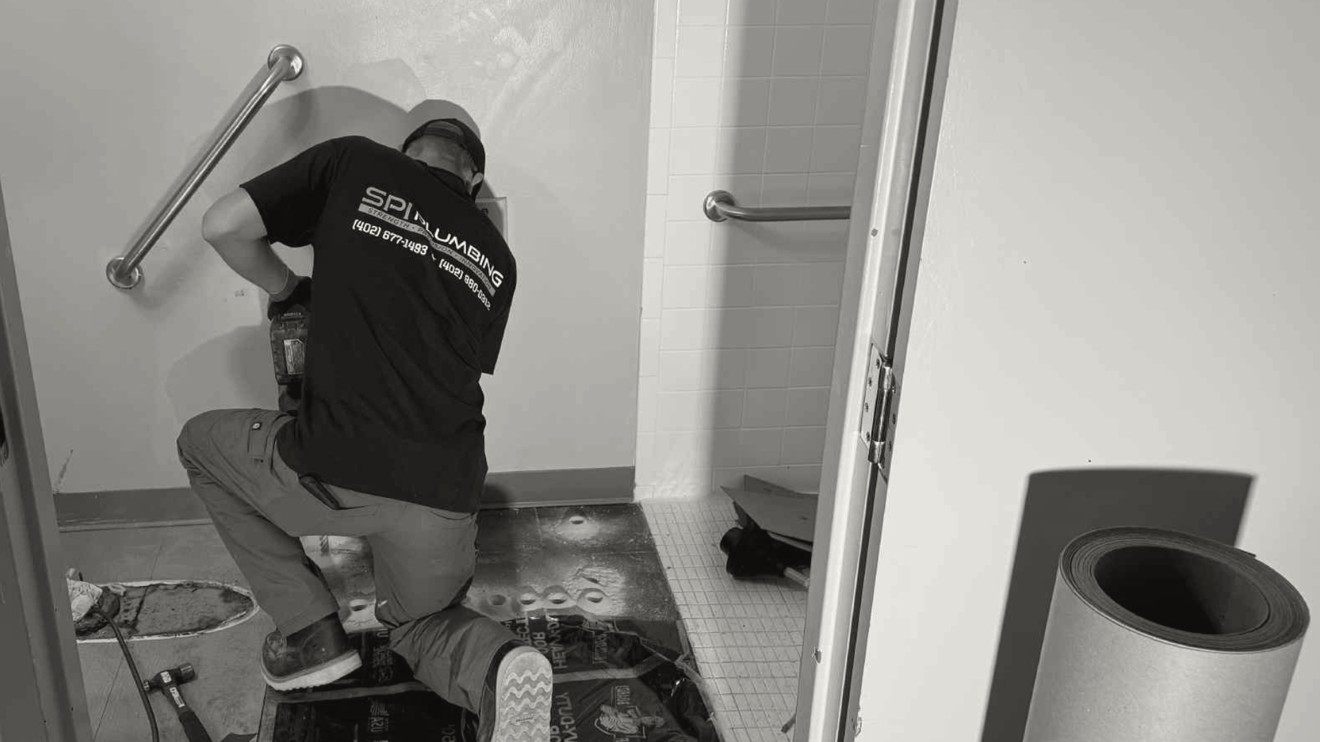 A worker kneeling inside a restroom, installing or repairing plumbing in a wall, wearing a company t-shirt and kneepads, with tools and construction materials around.