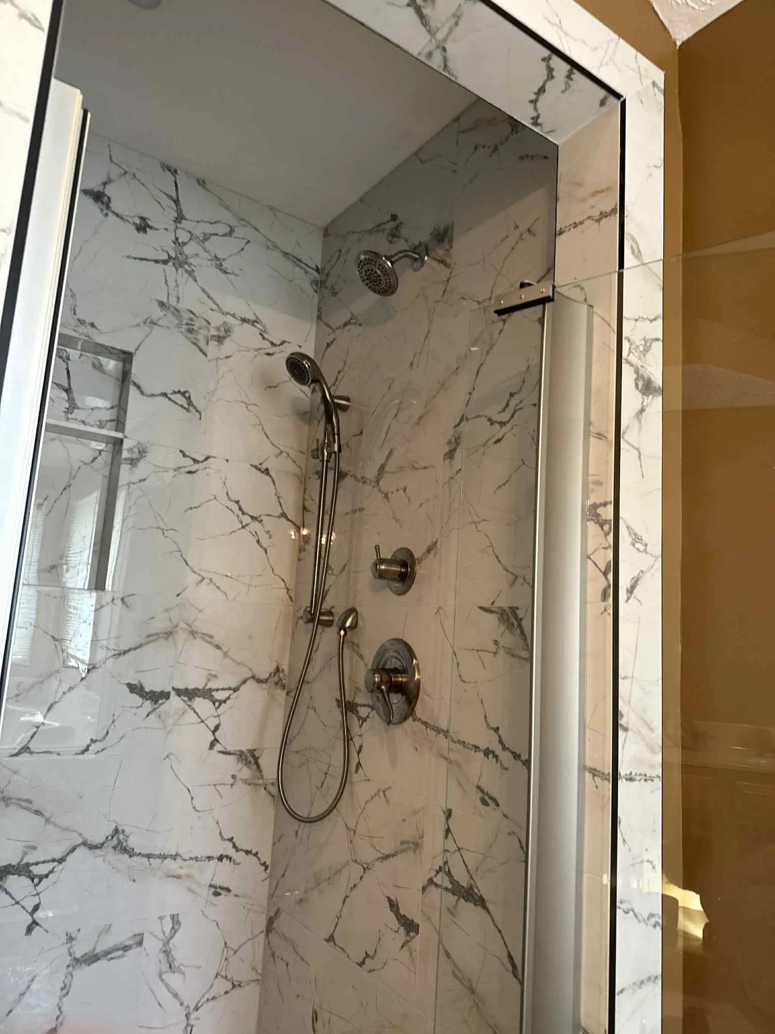 Shower with marble tiles, a handheld showerhead, a fixed showerhead, and a control knob, enclosed by a glass door.