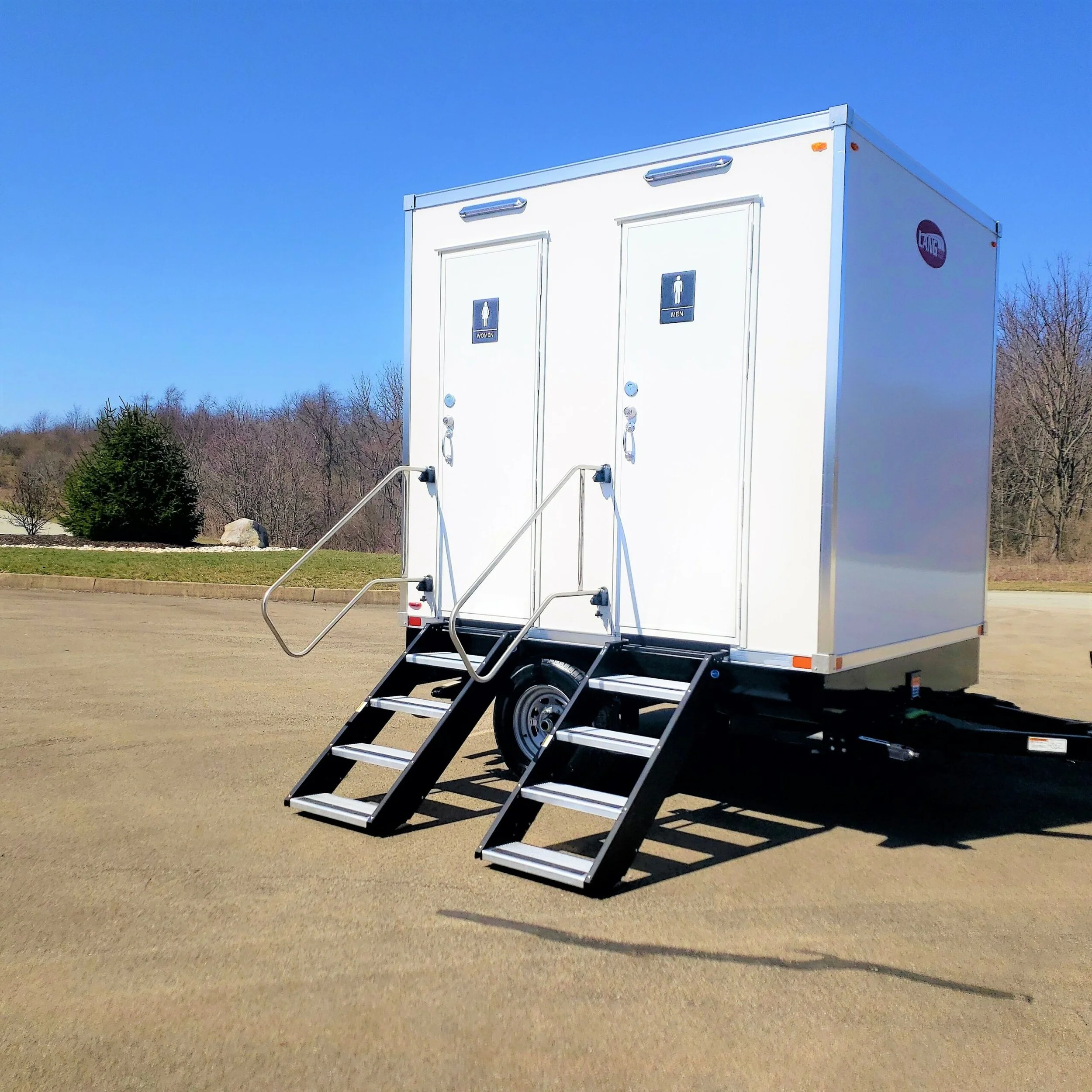 White portable restroom trailer with three steps and handrails leading to two separate bathrooms, set outdoors on a dirt lot with trees in the background.