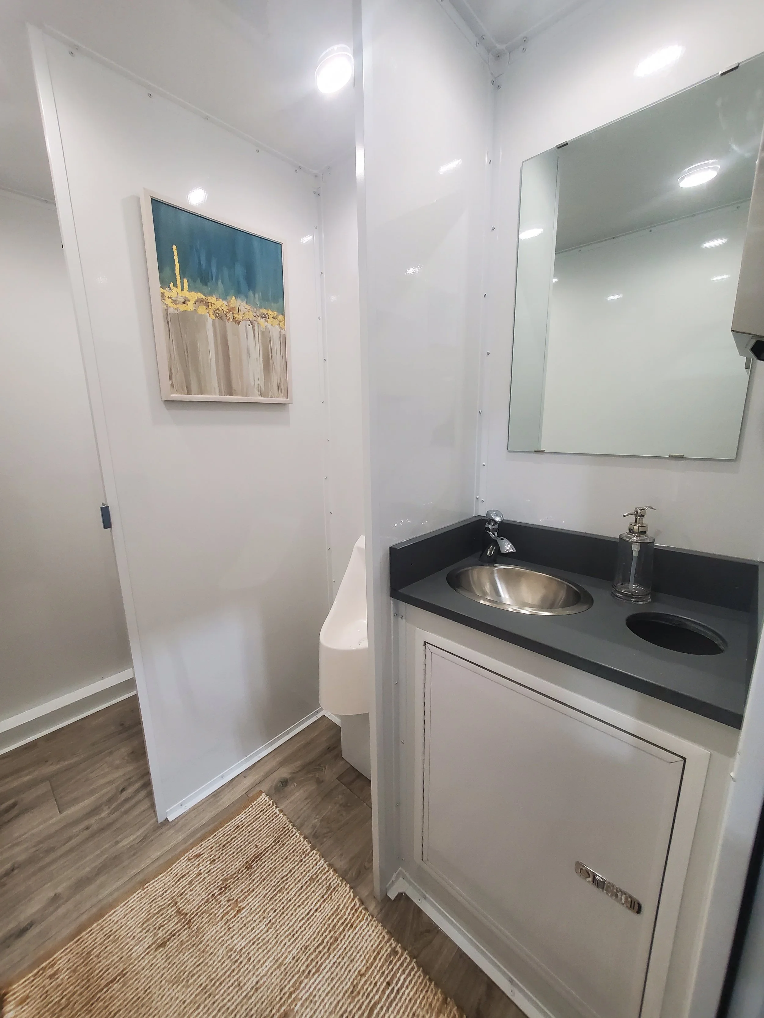 Small restroom with a sink and mirror, a urinal partially visible behind a partition, a windowed cabinet, and art hanging on the wall.
