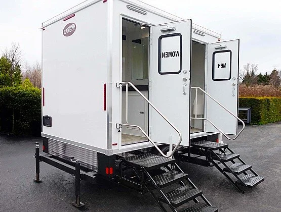 Mobile command or office trailer with stairs and two doors labeled 'WOMEN' and 'MEN', placed on an asphalt surface outdoors.