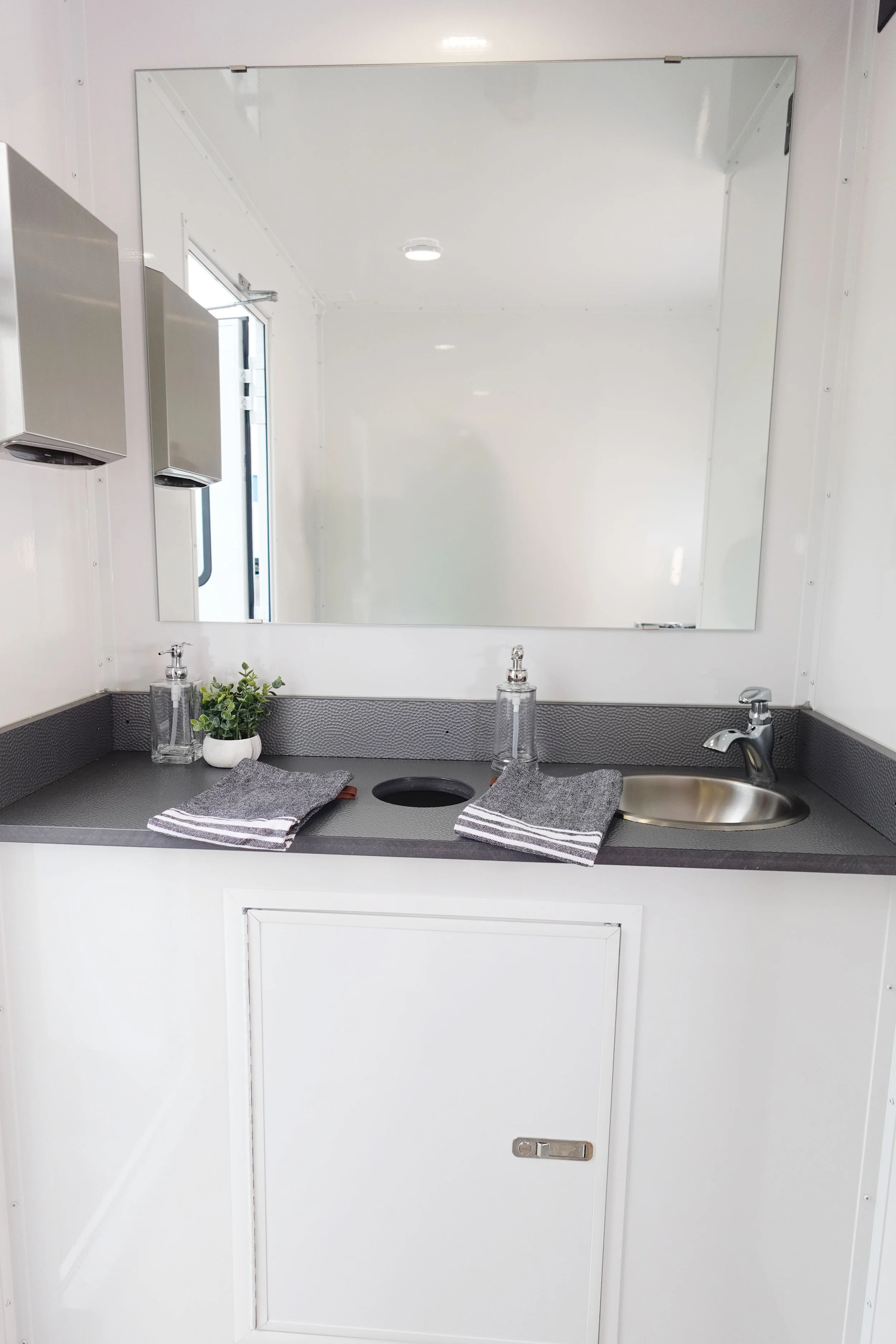 A small, clean sink area with a large mirror, two soap dispensers, a potted plant, and hand towels on a gray countertop.