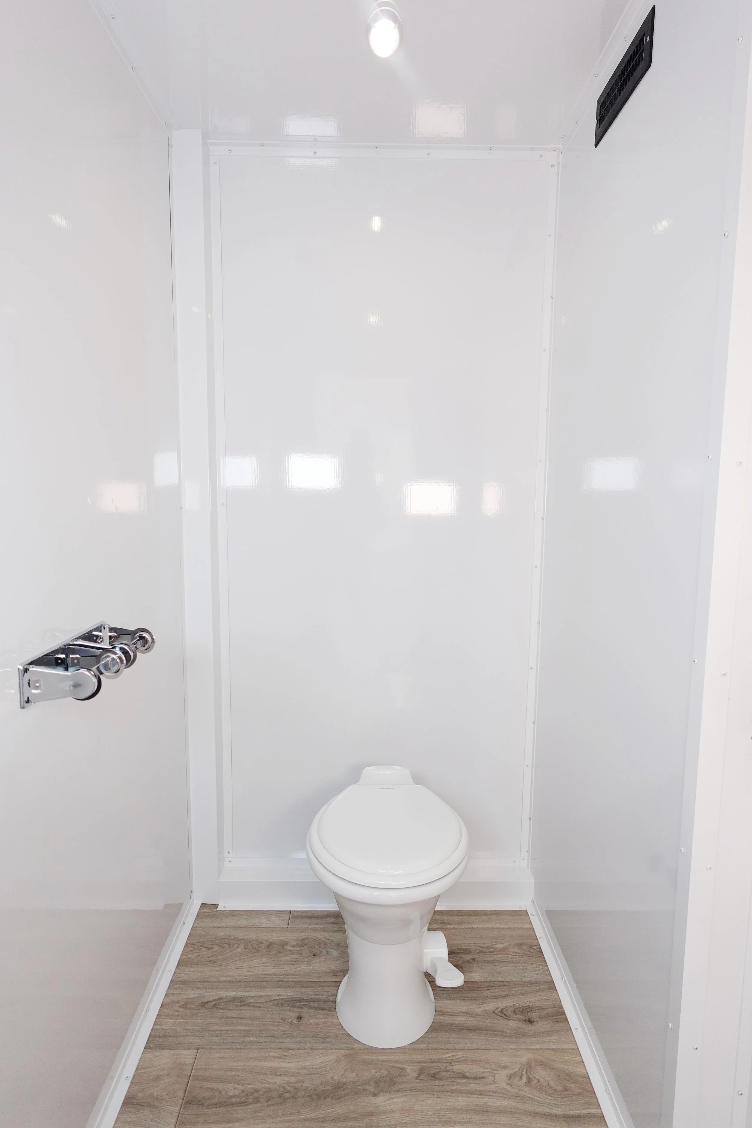 A small, minimalist bathroom with a toilet, a wood laminate floor, white walls, a wall vent, a metal pipe fixture, and ceiling lighting.