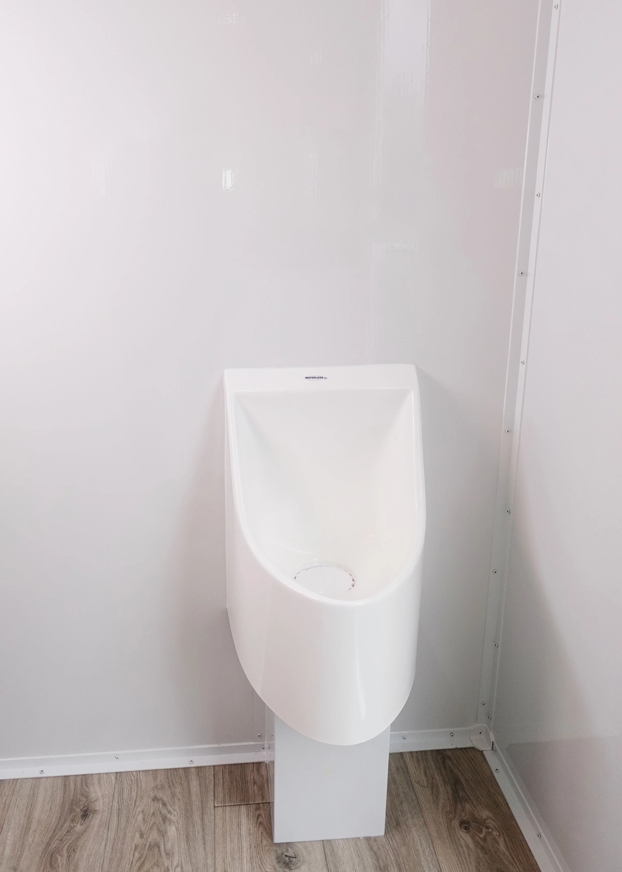 A white asymmetrical urinal mounted on a white wall in a restroom with wood flooring.