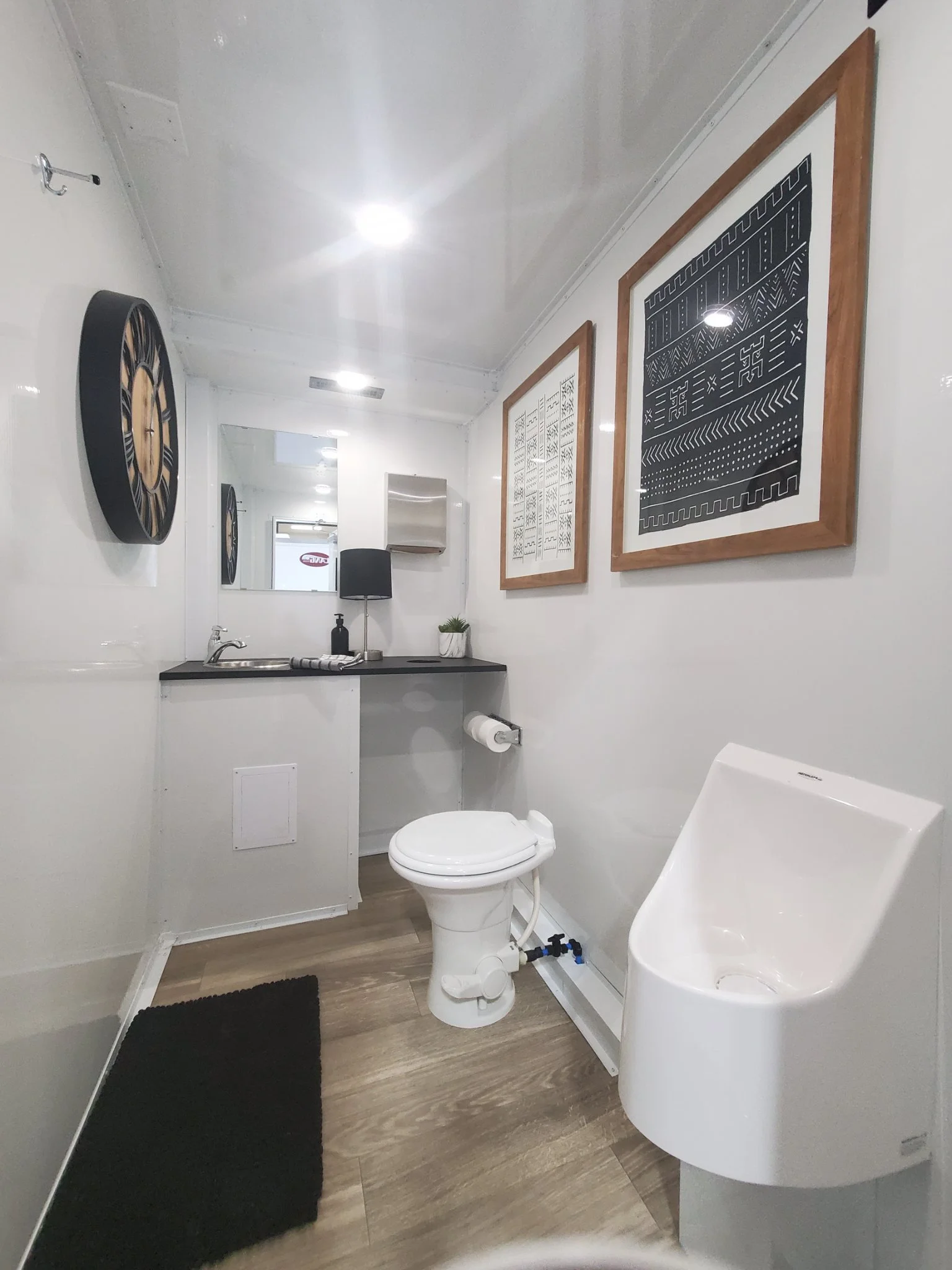 Small bathroom with white walls and wood floor, includes a toilet, a white urinal, a mirror, wall decorations, and a black rug.