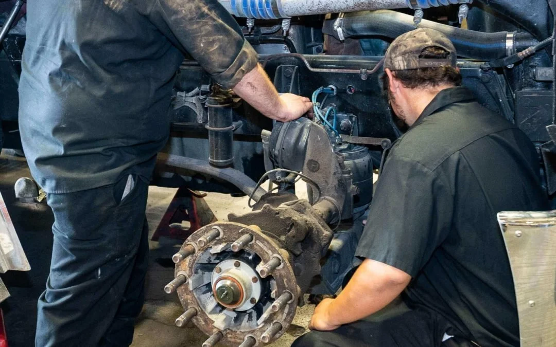 Two mechanics working on a large vehicle engine with the wheel hub removed, exposing the axle and brake components.