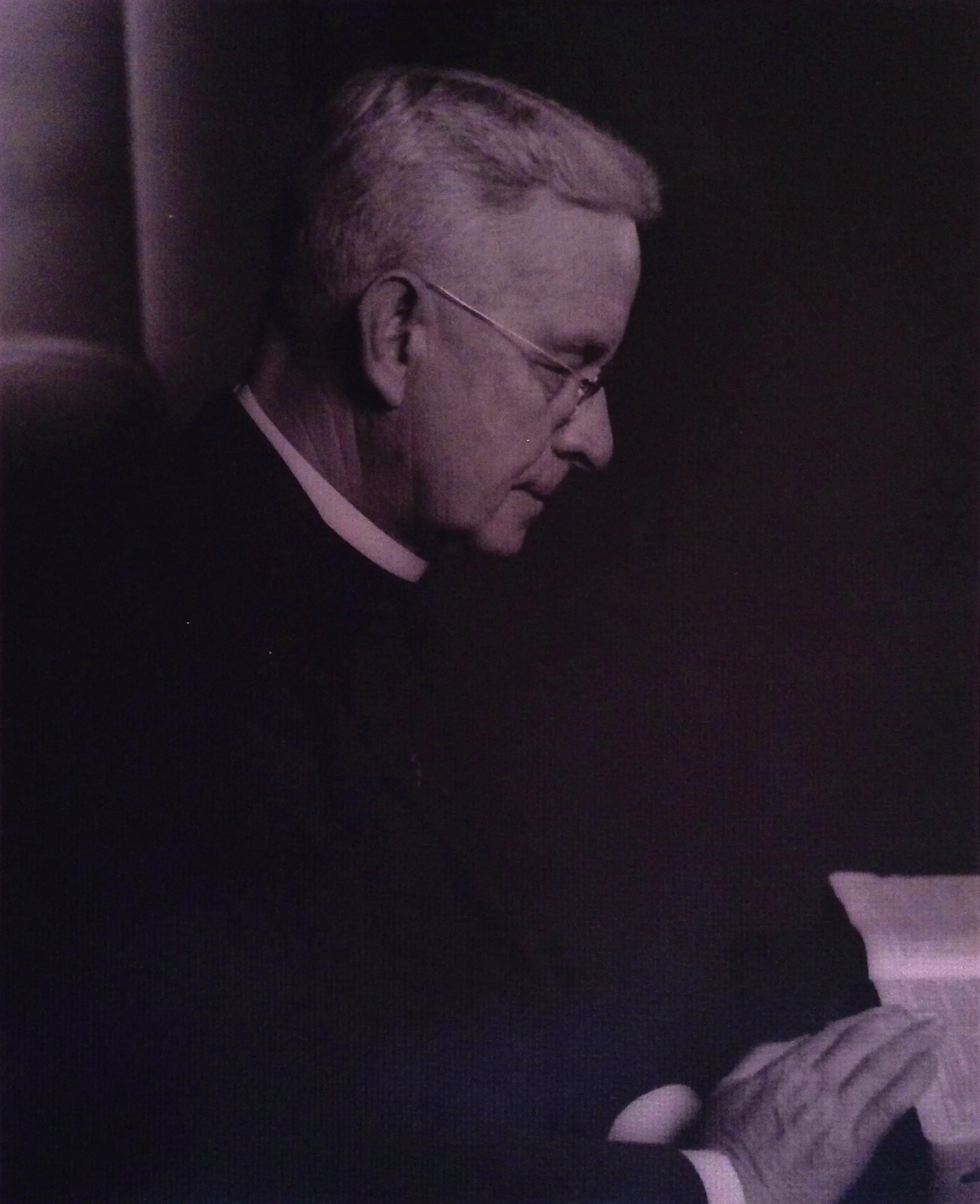 A black and white portrait of an older man with glasses, short light-colored hair, and a serious expression, looking downward.