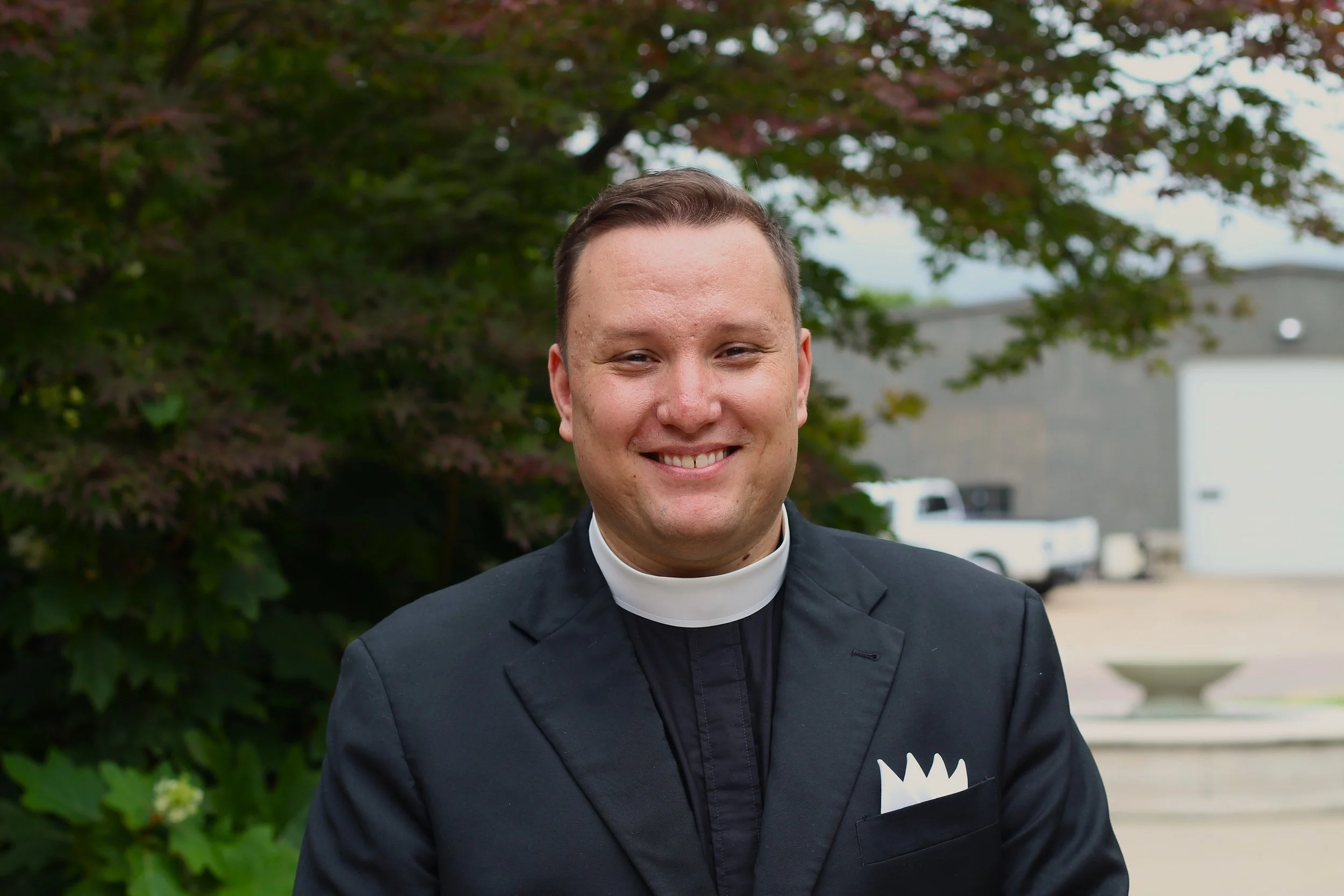 Portrait of a young man with short dark hair, wearing large clear glasses and a black clerical shirt with a white clerical collar, standing outdoors with blurred greenery in the background.