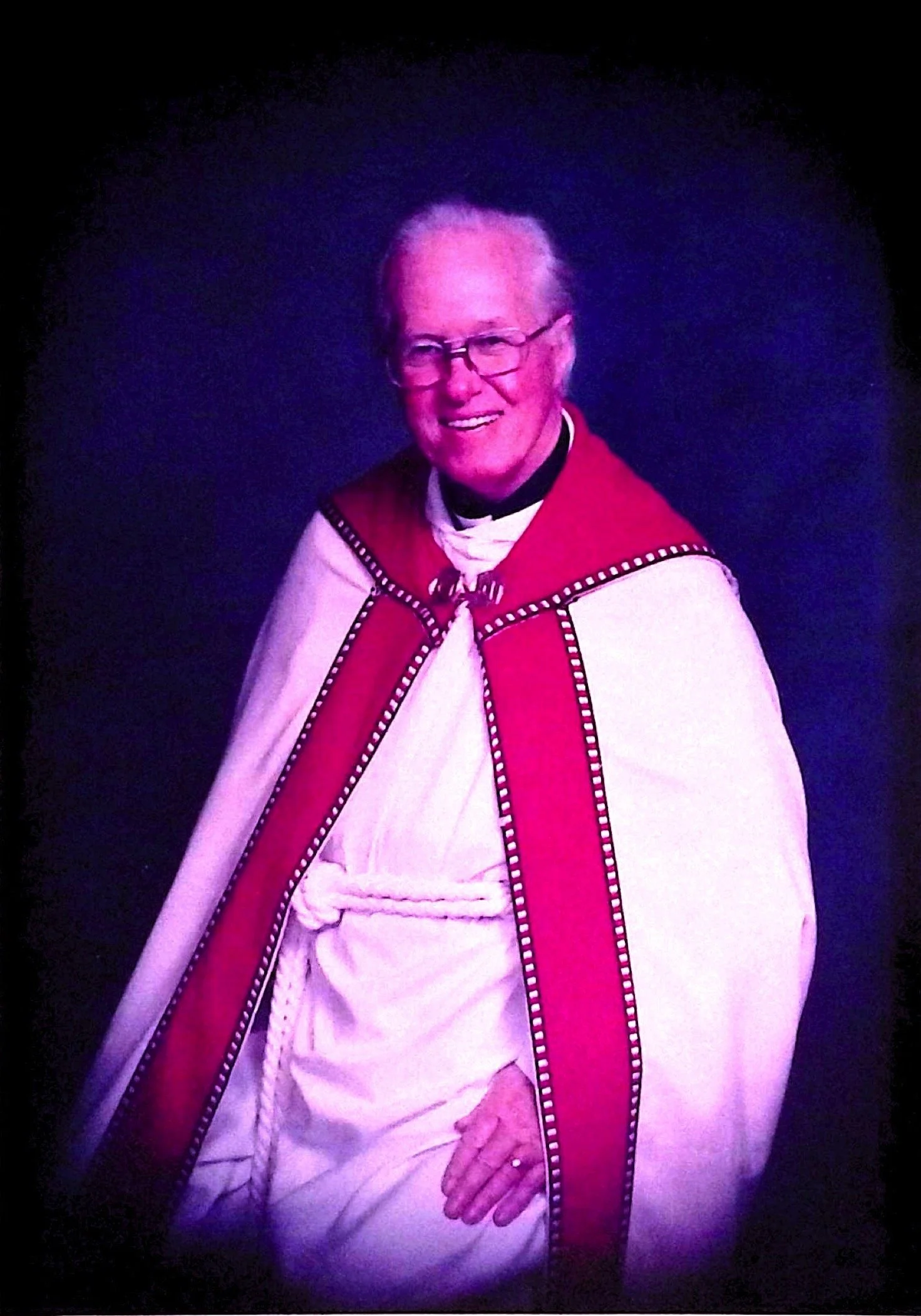A smiling elderly man dressed in religious robes, wearing glasses, with a white cloak and red ceremonial vestment featuring decorative trim.