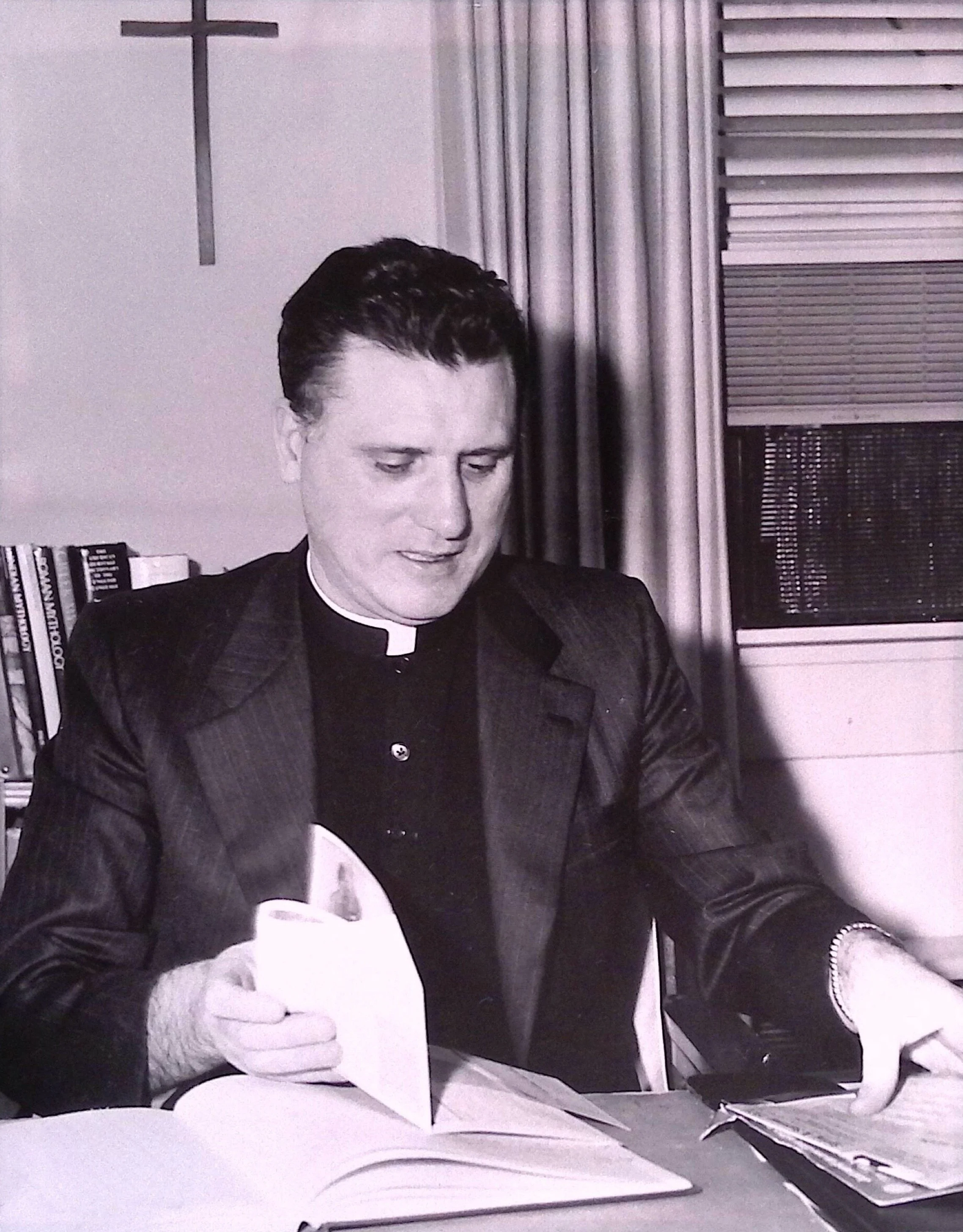 Black and white photo of a young man in a clerical shirt with a collar, sitting at a desk with open books in the background.