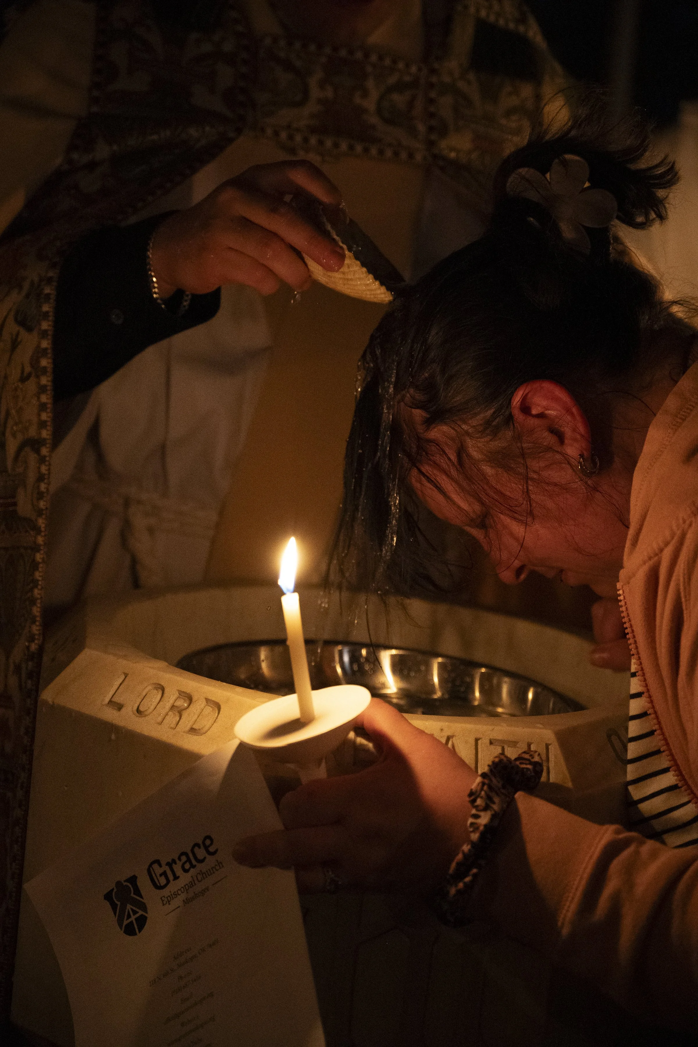 A woman during a baptism ceremony, with a person holding a candle and someone pouring water or oil over her head over a baptismal font at Grace Episcopal Church.