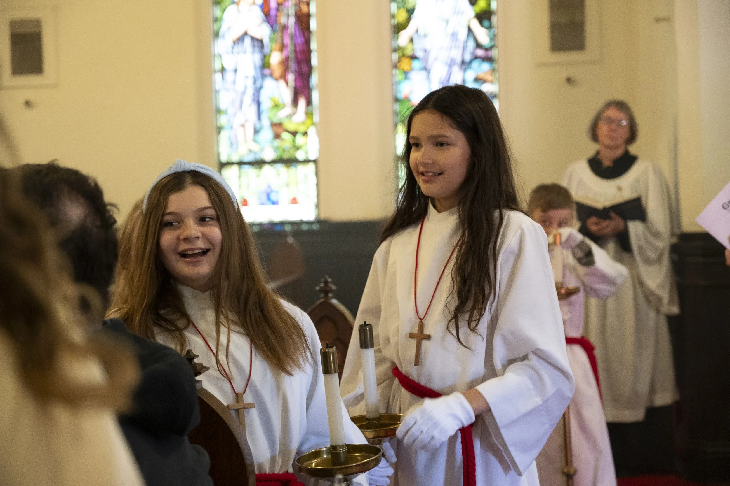 Children dressed in white robes with crosses on necklaces, participating in a church ceremony, with stained glass windows in the background.