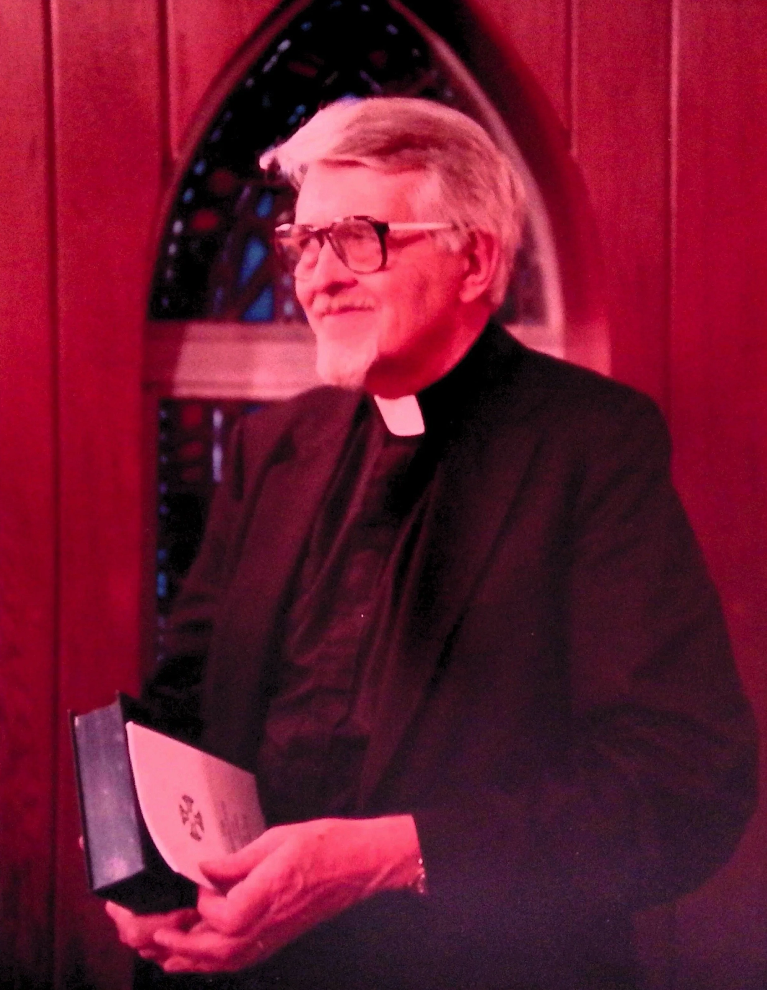 A person dressed as a priest, wearing glasses and a clerical collar, covering their face with one hand. The background appears to be a decorative or architectural element.