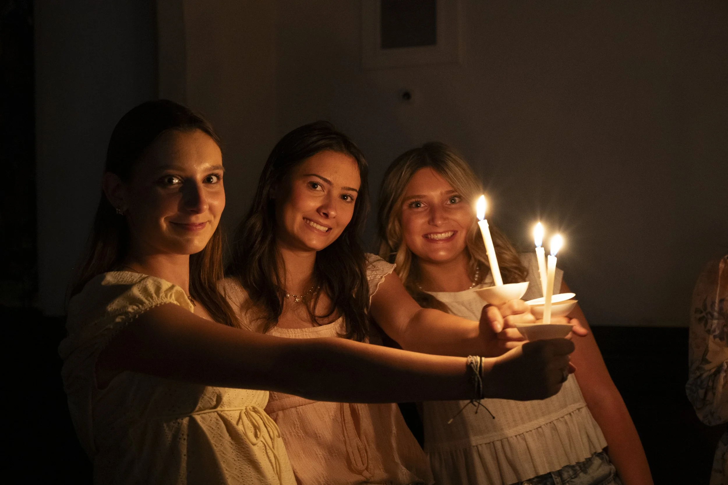 Three women smiling in a dark setting holding lit candles in small bowls, celebrating together.