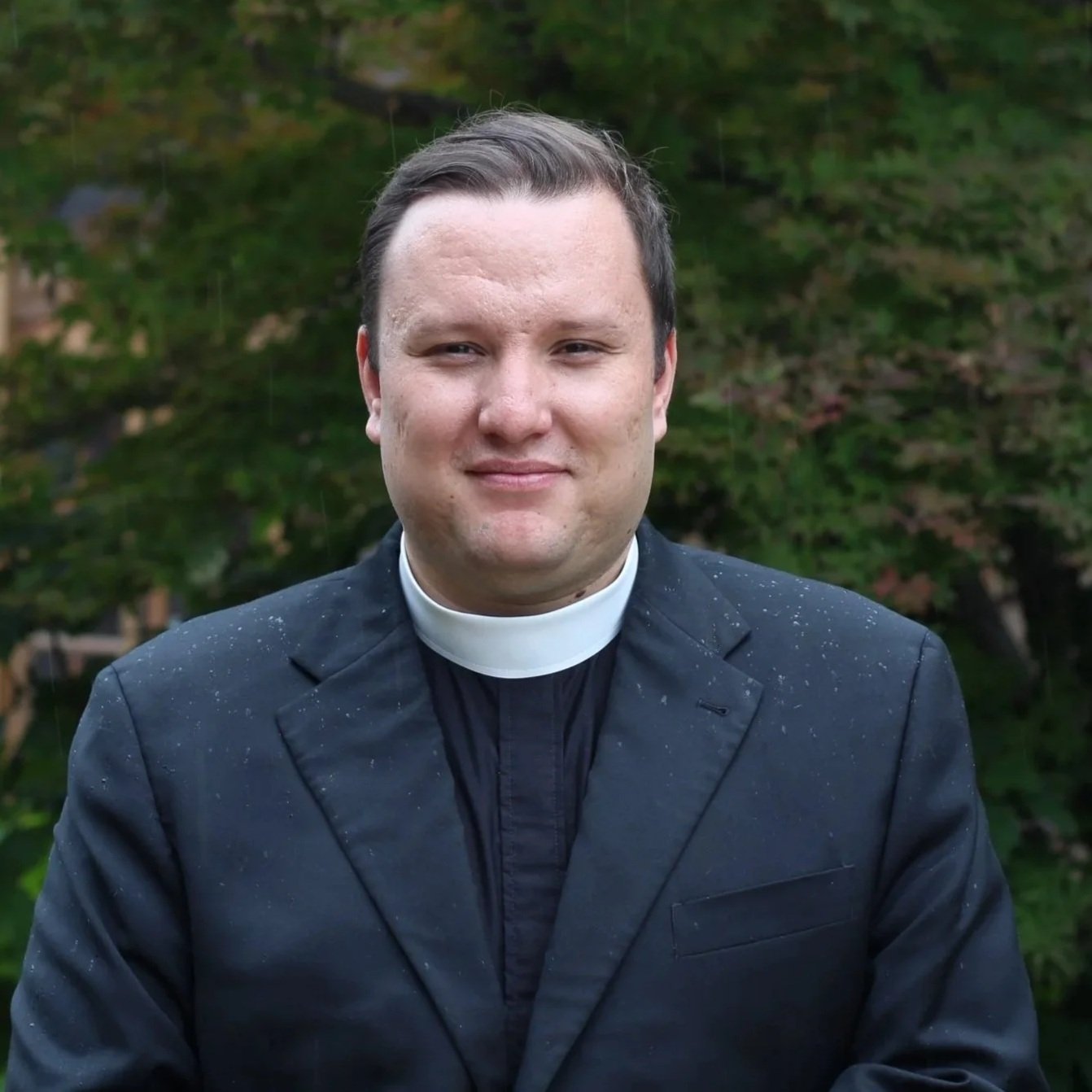 Portrait of a young man with short dark hair, wearing large clear glasses and a black clerical shirt with a white clerical collar, standing outdoors with blurred greenery in the background.