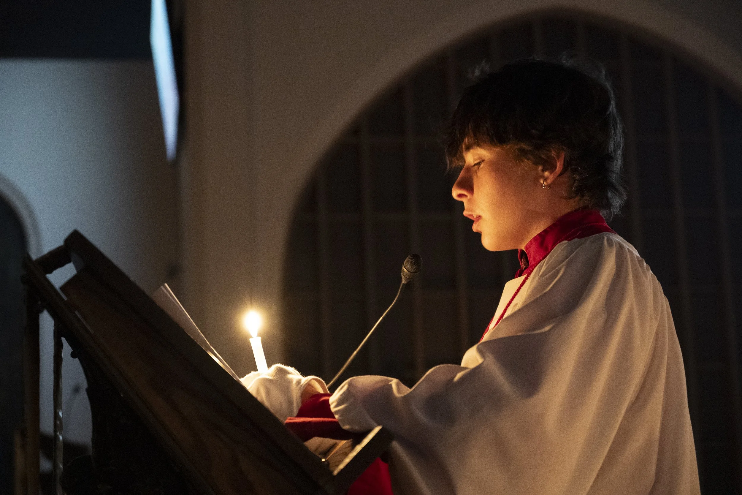 A young person with short dark hair reading or singing from a music stand while playing the piano in a dimly lit church, wearing a white robe with a red collar.
