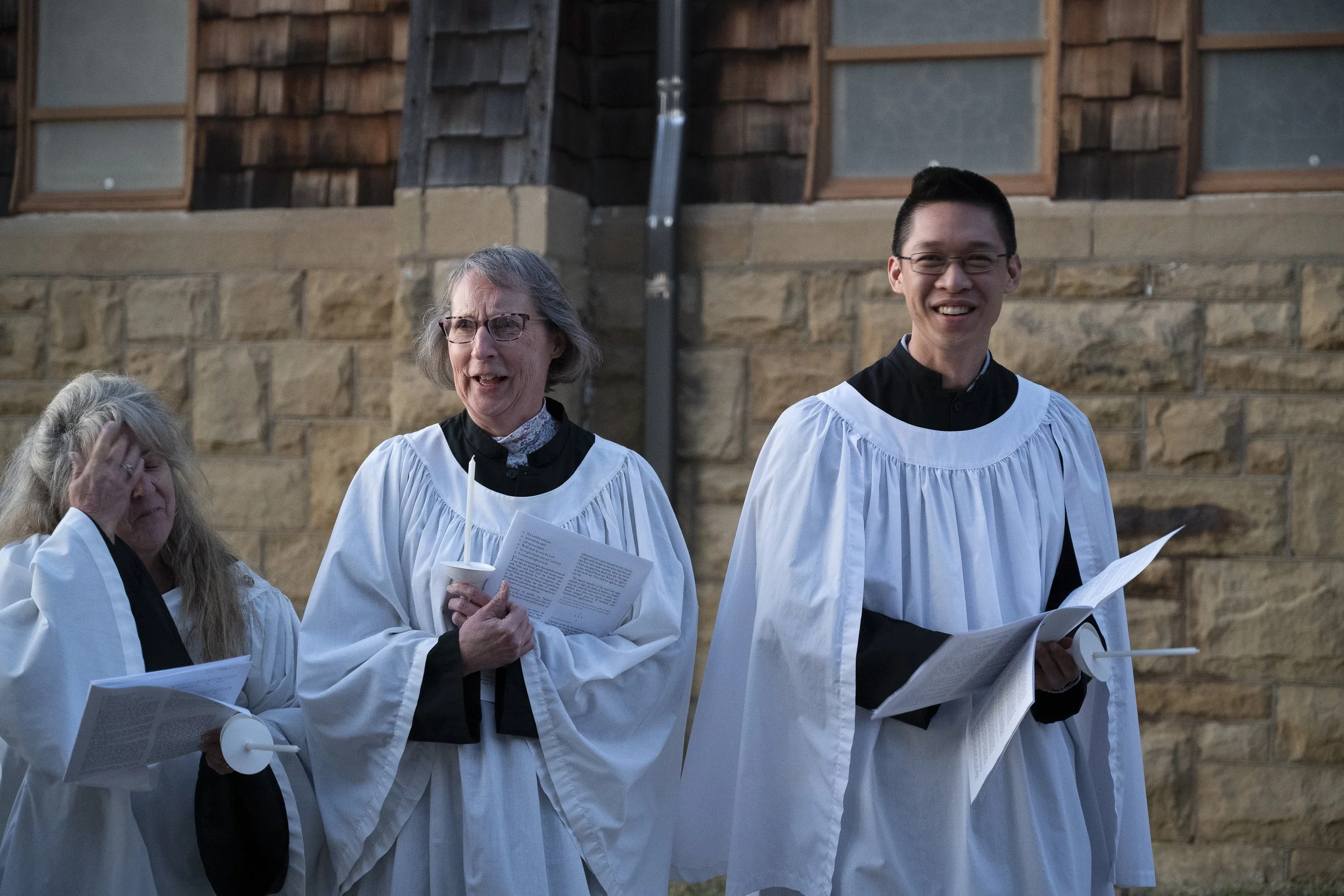 Three people dressed in choir robes standing outdoors in front of a stone wall, holding papers and candles, with two women on the left and a man on the right.