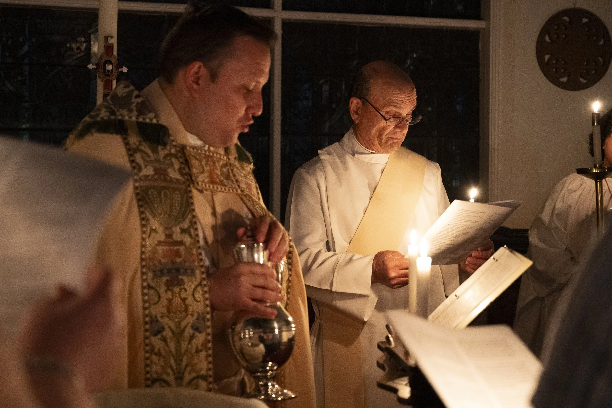 Two clergy members standing in a dimly lit room, reading from liturgical books illuminated by candles during a religious ceremony.