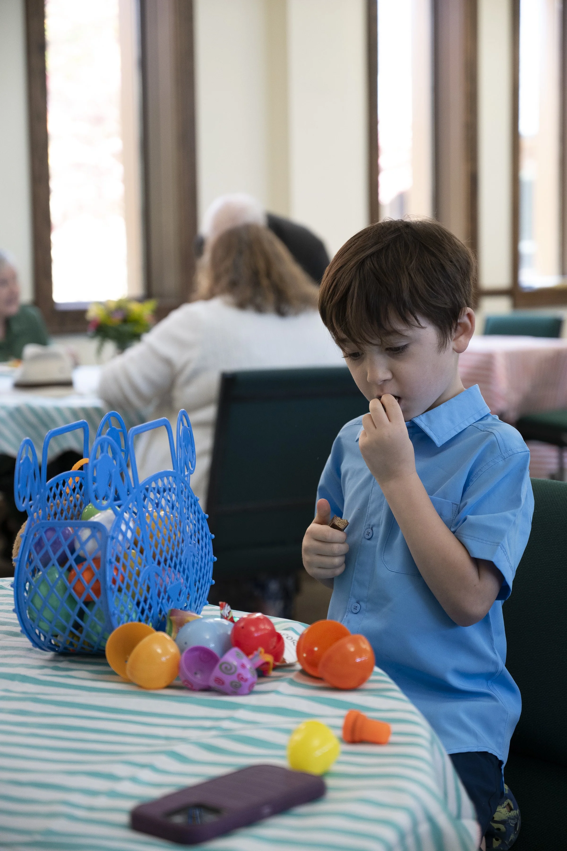 A young boy in a blue shirt examines toy Easter eggs on a table during a gathering in a bright, indoor setting.