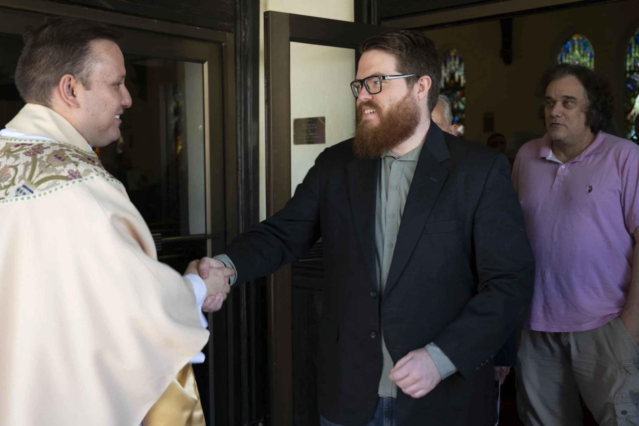Two men shaking hands at the entrance of a church with stained glass windows, while a third man in a pink shirt looks on.