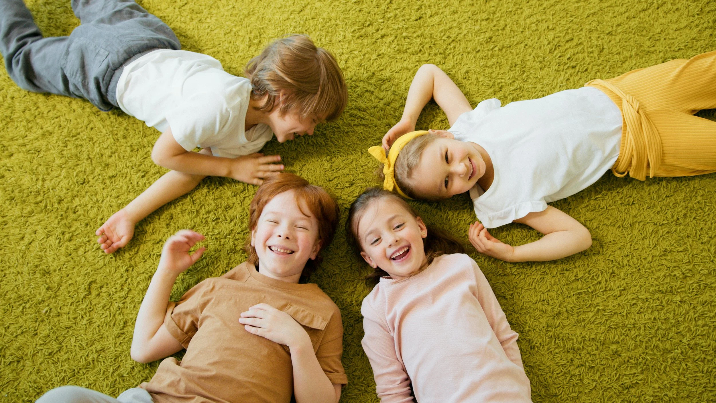 Five smiling children lying on a yellow carpet, facing the camera, engaging in playful interaction.