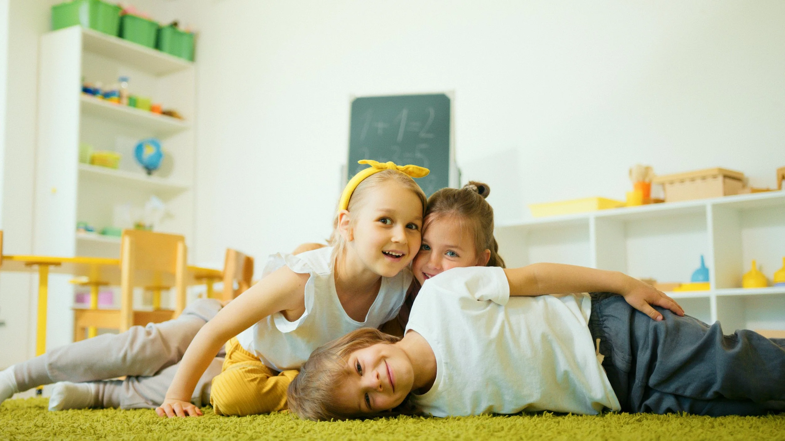 Three children playing and lying on a green rug in a classroom, smiling and cuddling together.