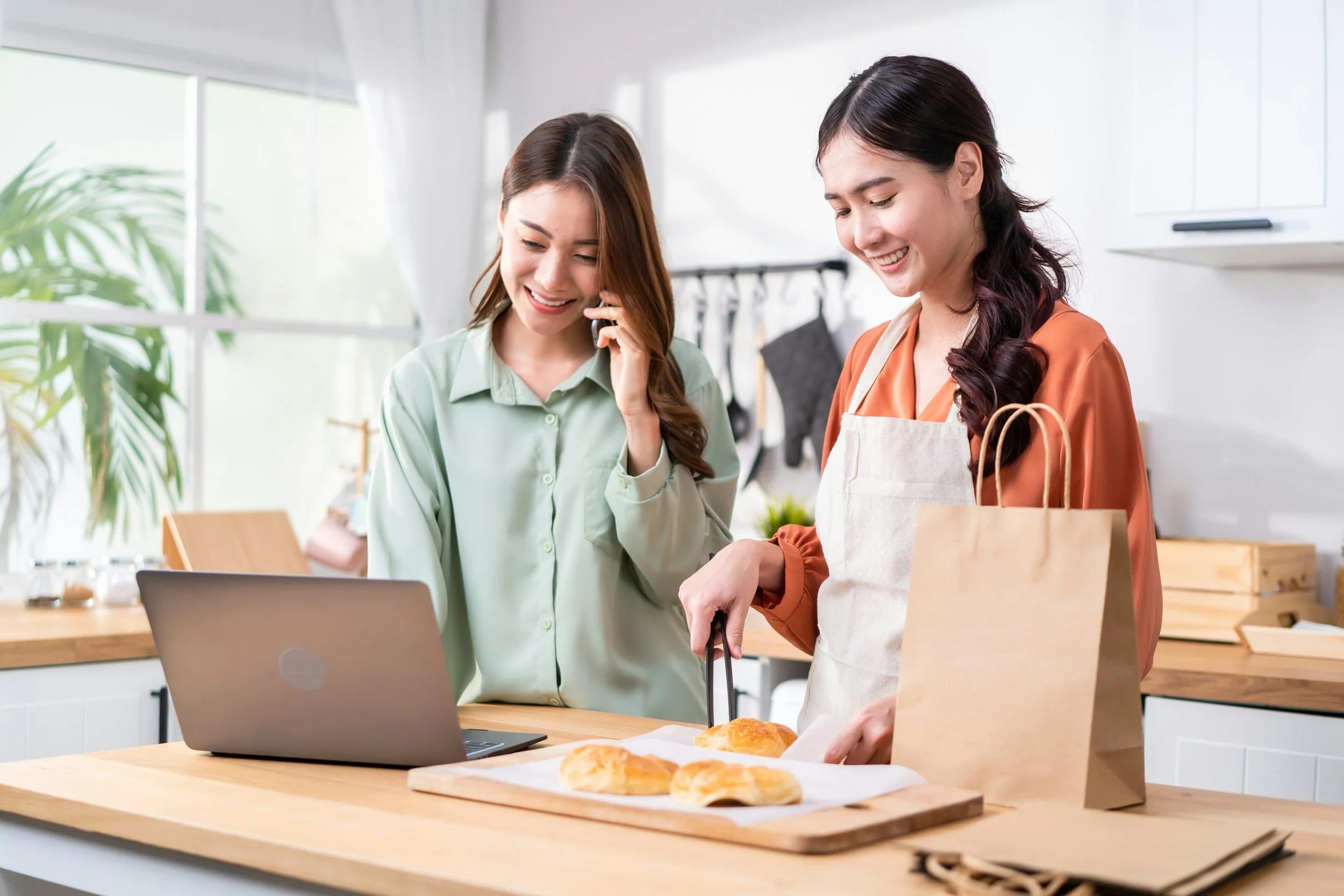 Two women preparing baked goods in a modern kitchen, one slicing bread and the other talking on a cell phone, with a laptop, paper bags, and baked bread on the counter.