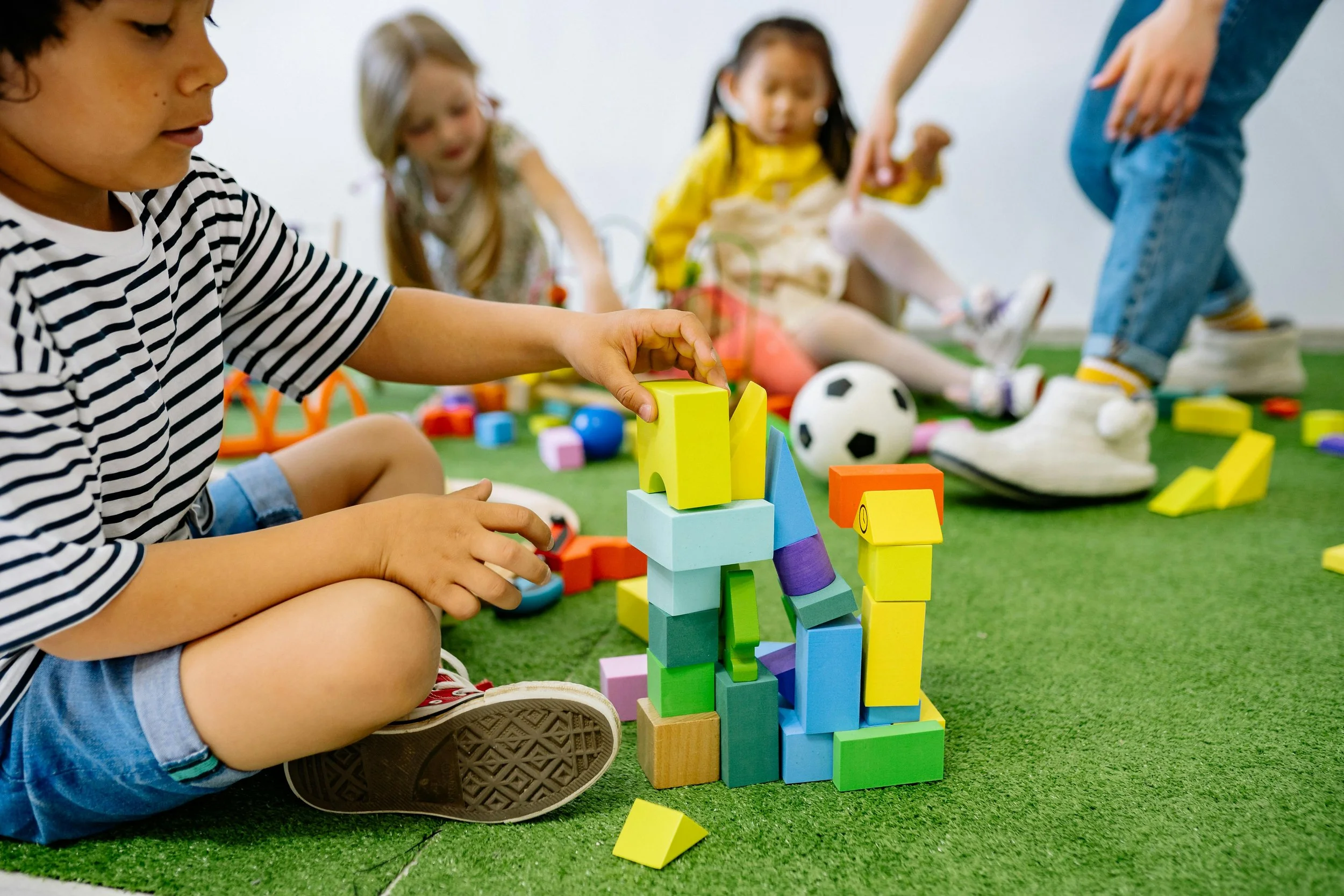 Children playing with colorful building blocks on a green carpet, with a soccer ball and scattered toys in the background.