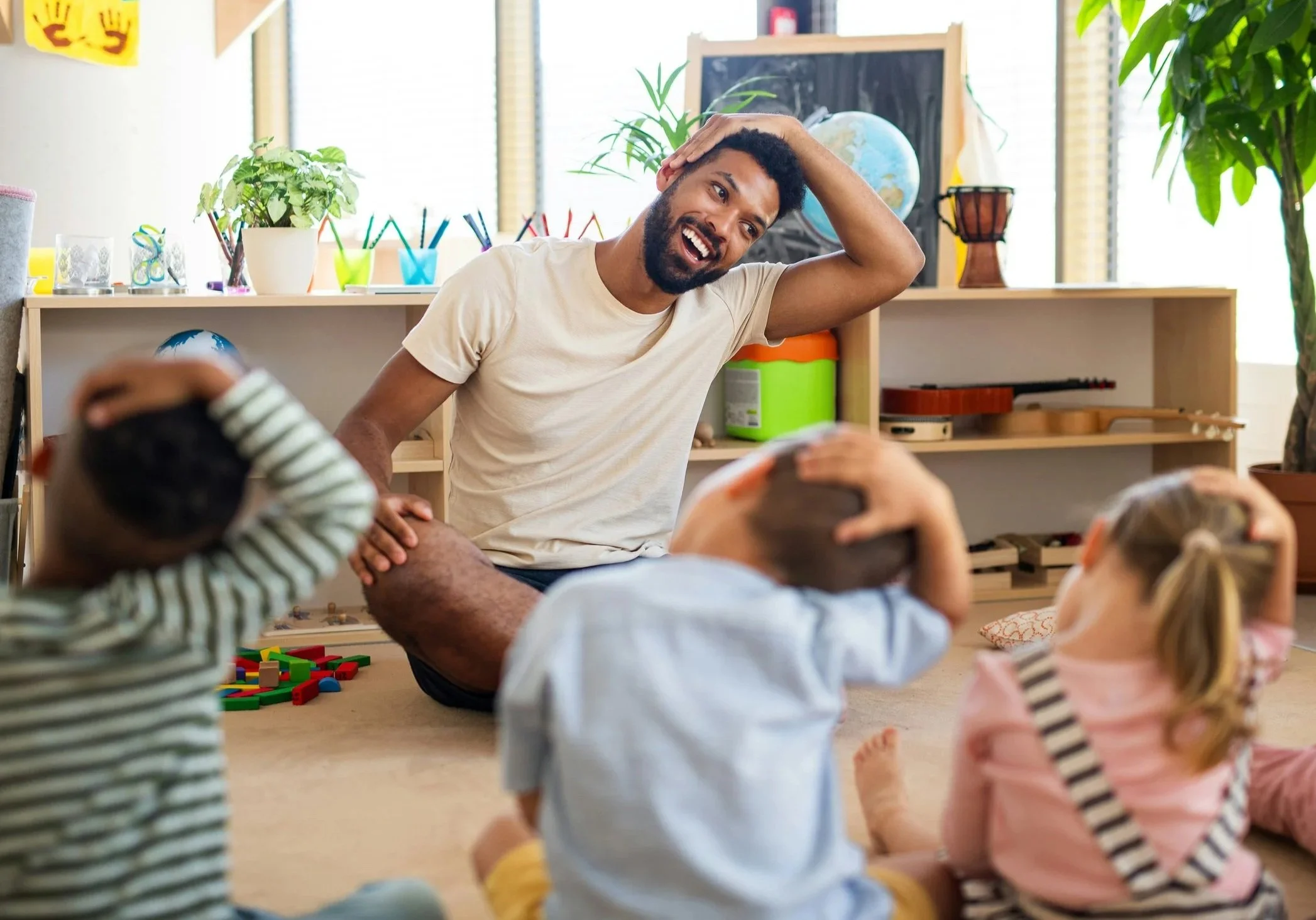 A man leading a group of young children in a song or activity in a classroom, sitting on the floor with children around him, some with hands on their heads, smiling and engaging, with colorful classroom decorations and plants in the background.