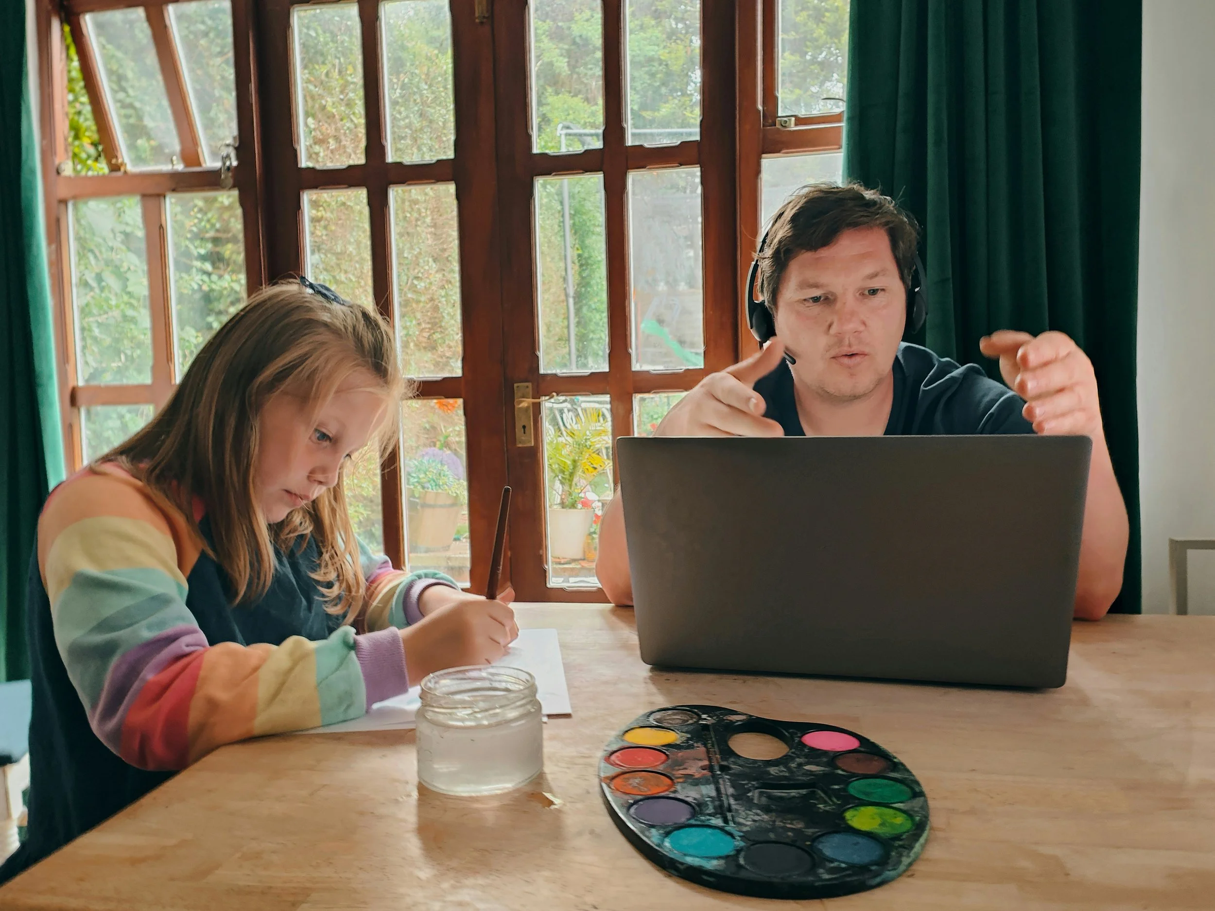 A young girl with long hair, wearing a colorful striped sweater, is drawing with a pencil at a wooden table. An adult man with short dark hair, wearing a black shirt and headphones, is sitting next to her and working on a laptop. There are watercolor paints and a jar of water on the table. The background shows large window panes with a view of greenery outside.