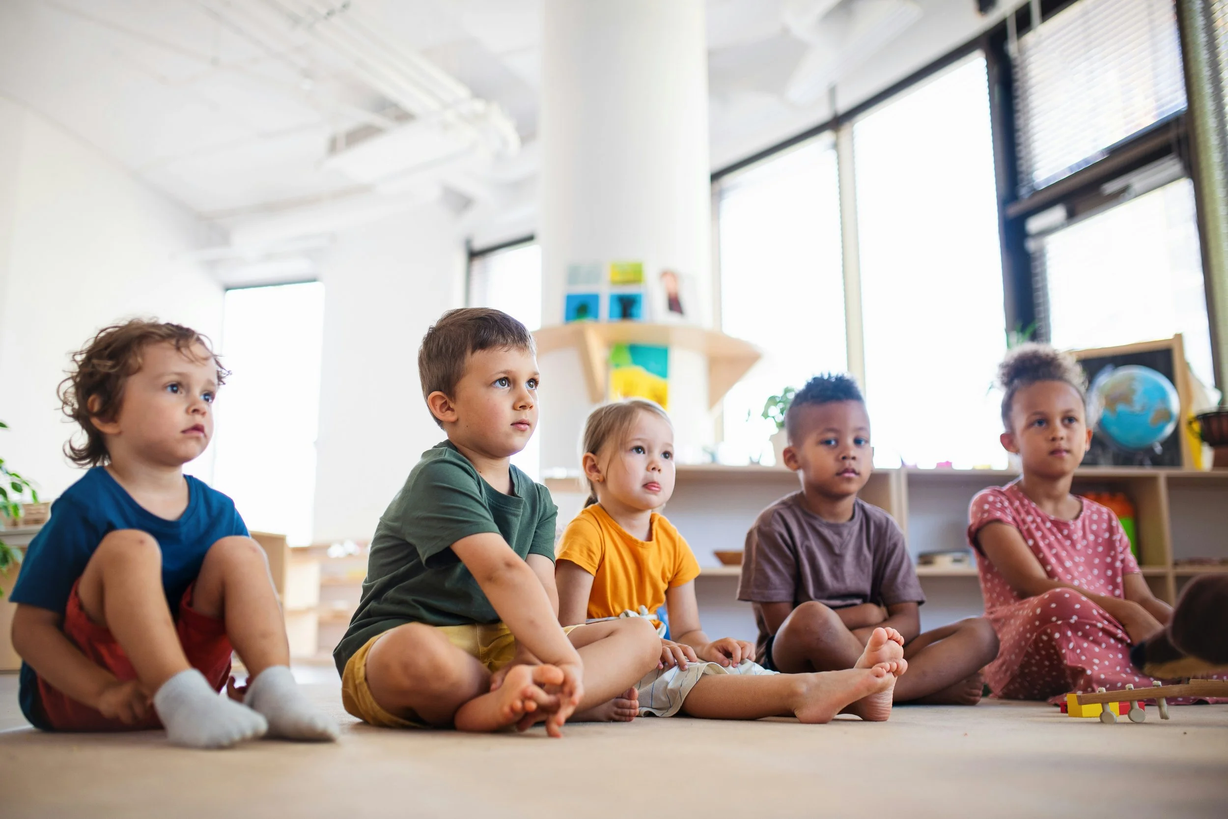 Five children sitting on the floor listening attentively in a classroom.