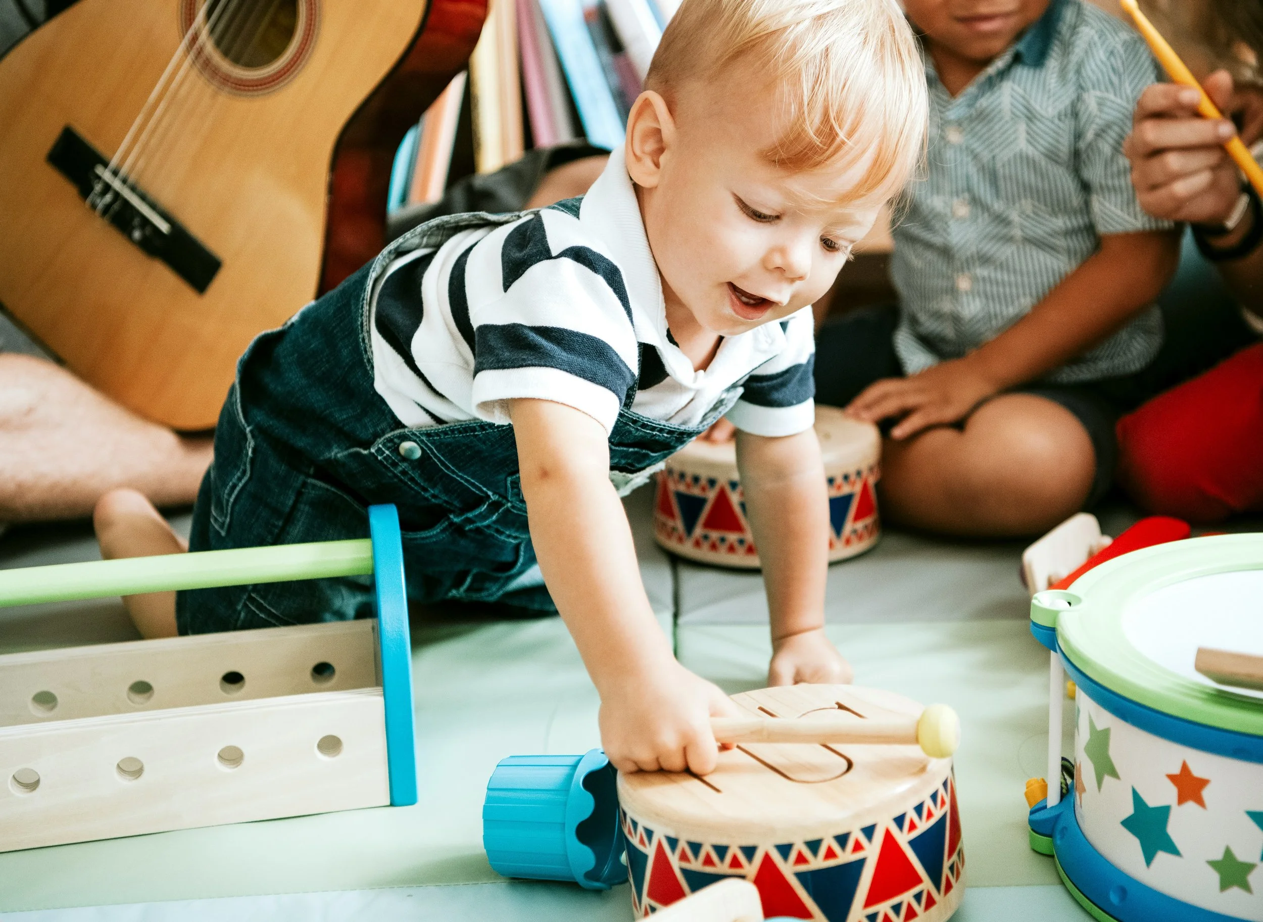Young boy with blonde hair playing with a wooden toy on the floor surrounded by musical instruments and toys.