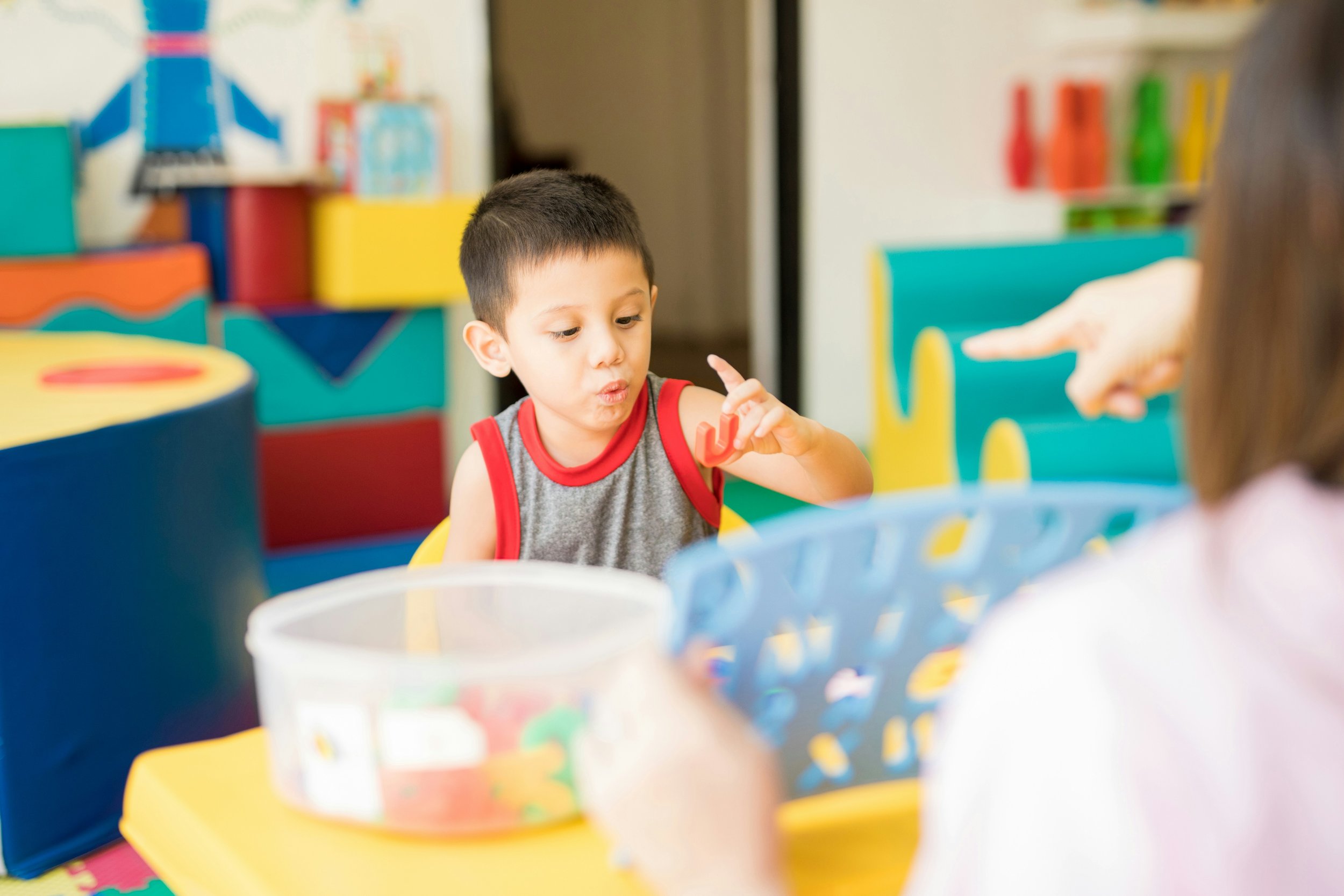 A young boy with short dark hair wearing a sleeveless gray shirt with red trim, looking at a red toy while sitting at a colorful table in a child play area. A girl with long hair holding a basket is visible in the foreground. Background contains bright, multicolored blocks and toys.