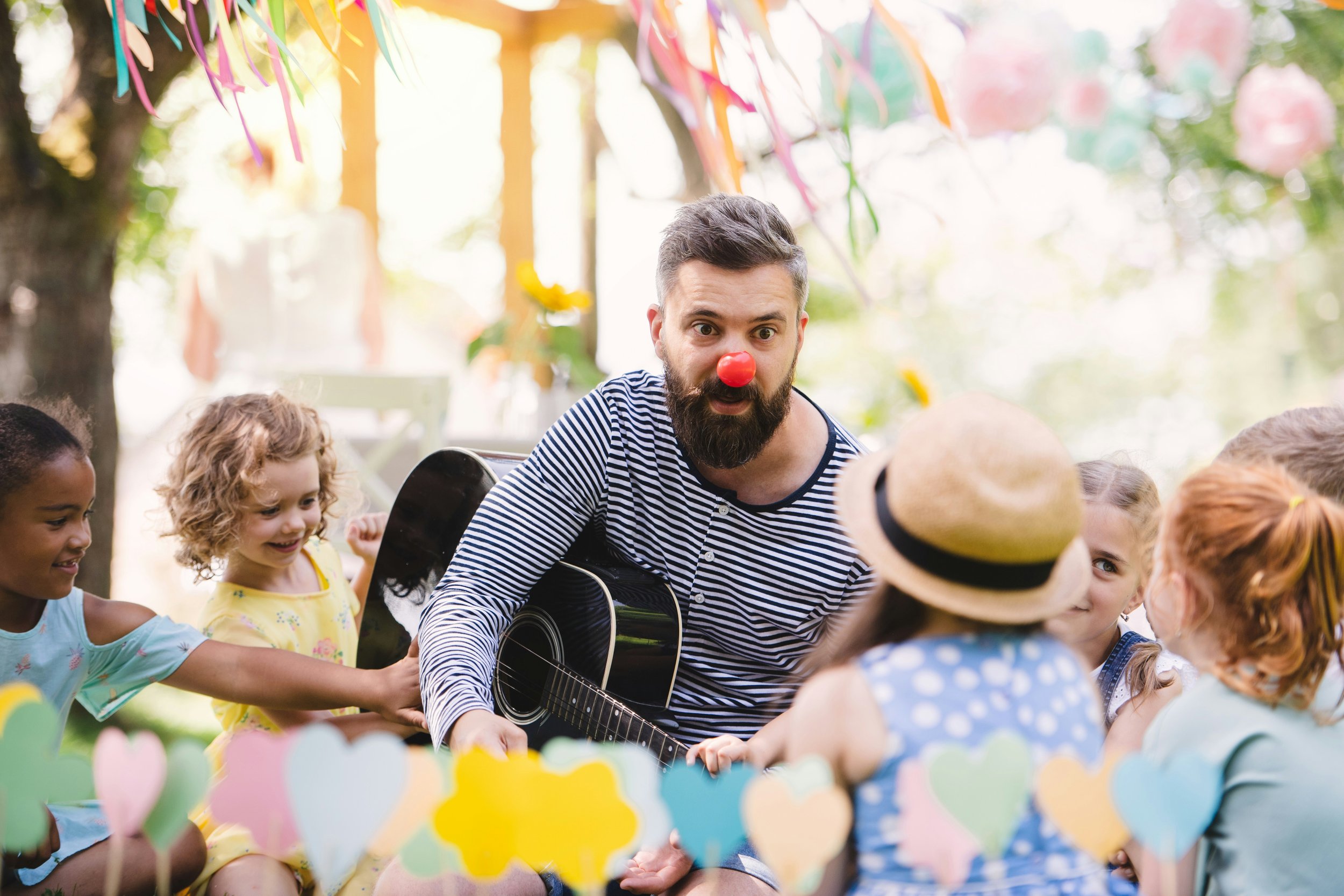 A man with a red clown nose and a striped shirt plays guitar while children sit around him, enjoying a colorful outdoor party with decorations and smiling faces.