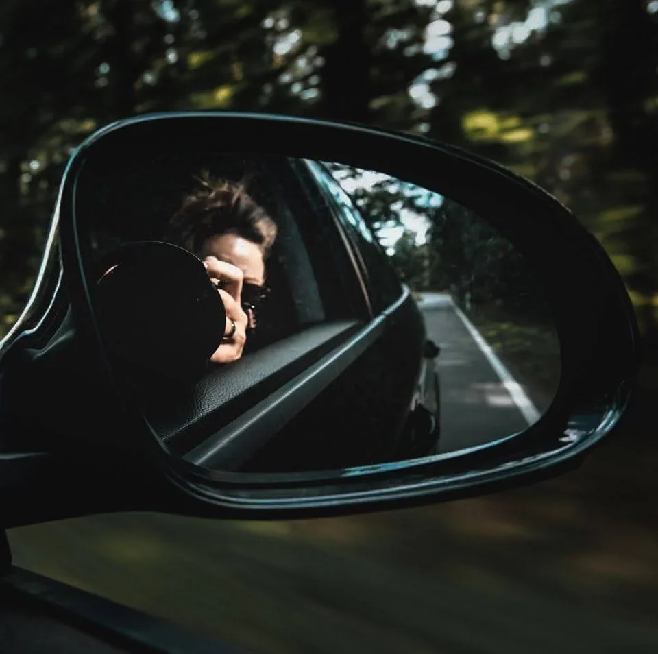 Eine Frau mit Kamera spiegelt sich im Autospiegel, während die Straße hinter ihr verschwimmt – Momentaufnahme zwischen Bewegung und Selbstblick.