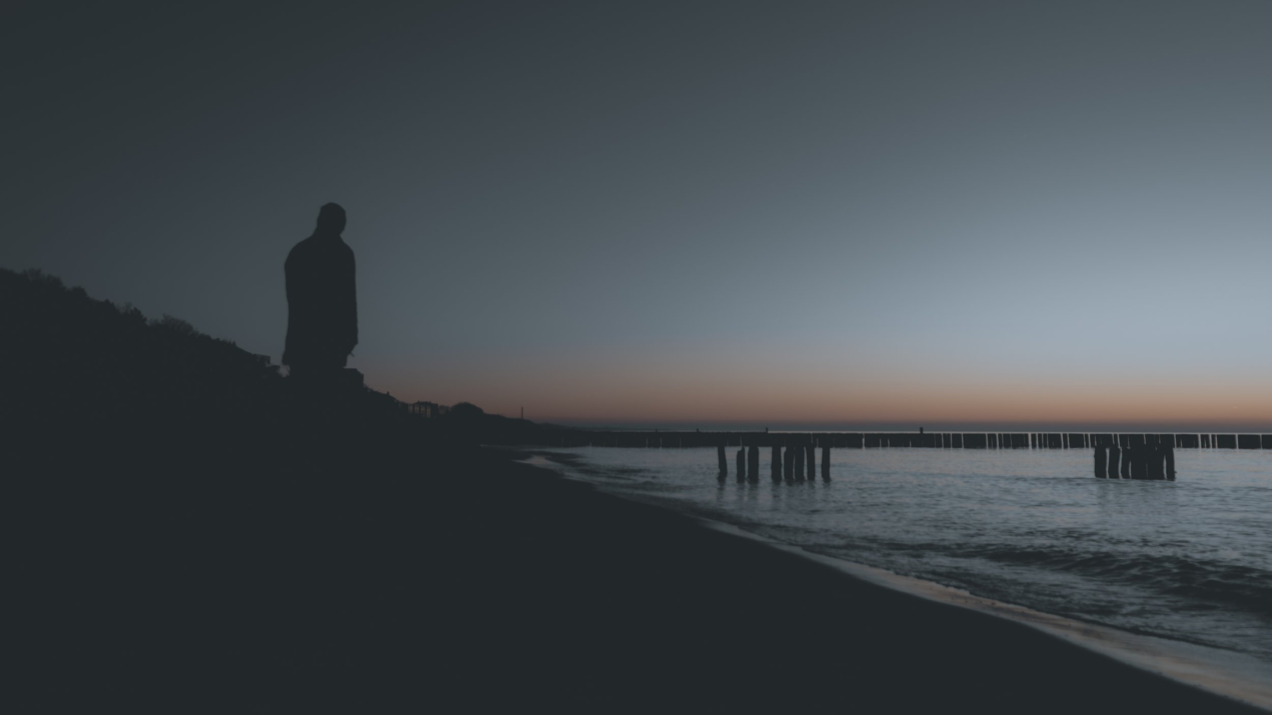 Am Rand
Eine einzelne Silhouette steht zwischen dunkler Küste und dem letzten Licht über dem Meer. Mensch und Landschaft bleiben auf wenige Formen und einen schmalen Horizont reduziert.