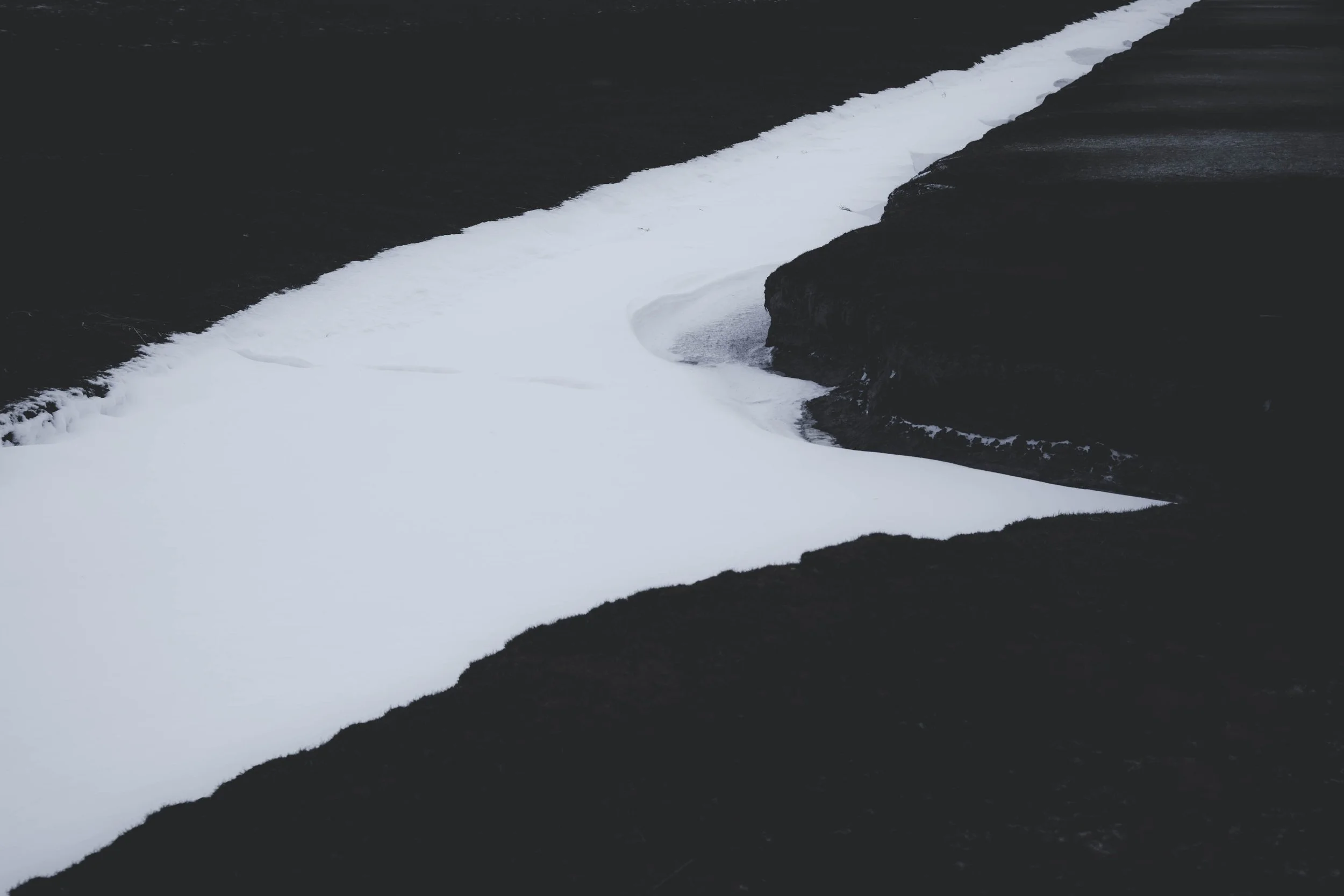 Weiße Spur
Eine geschwungene Schneefläche durchschneidet das dunkle Küstenland. Die starke Reduktion löst die Landschaft beinahe in eine grafische Form auf.