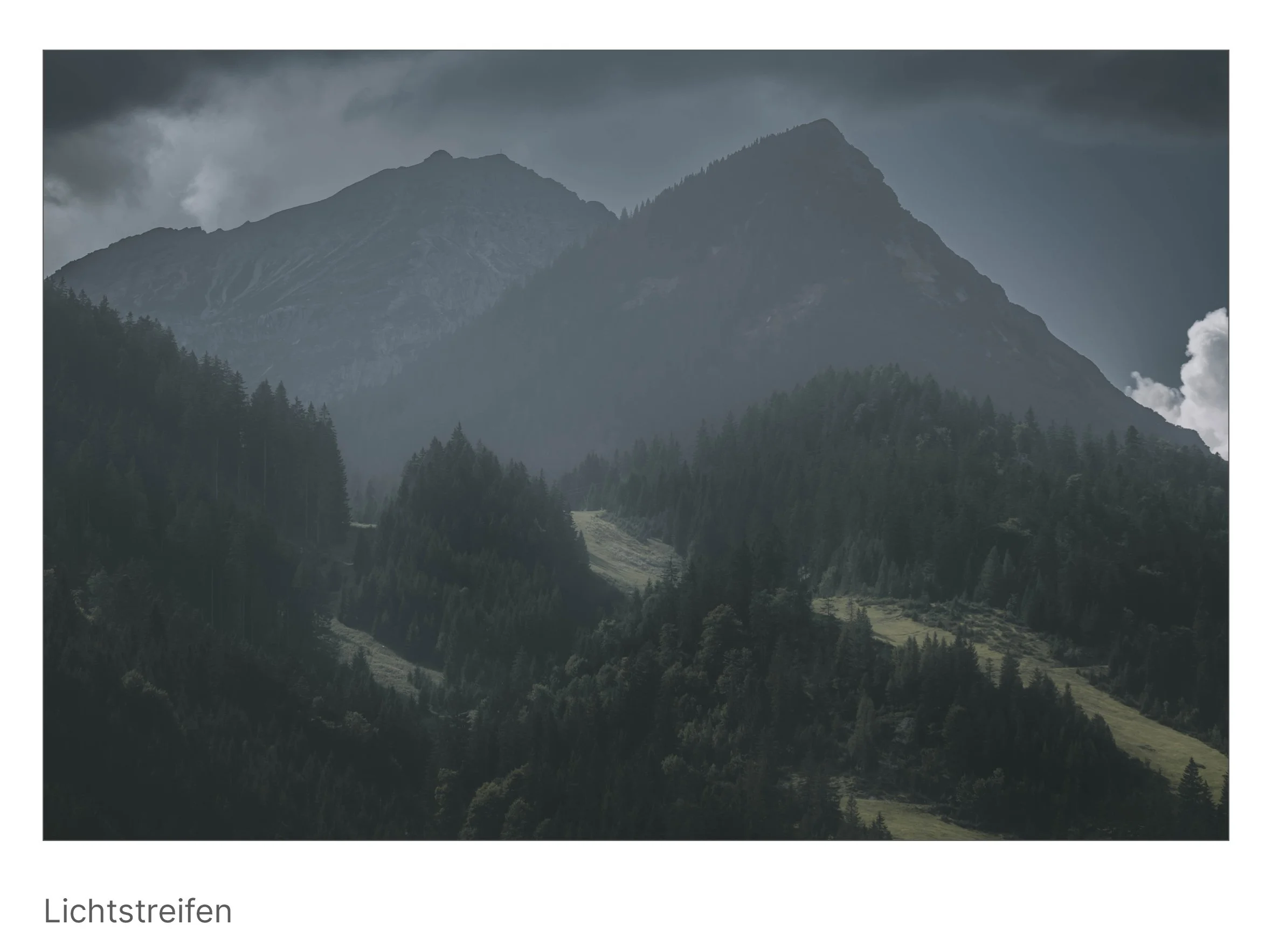 Ein heller Streifen Sonne durchbricht dunkle Wolken über bewaldeten Hängen – Hoffnungsschimmer in gedämpfter Landschaft.