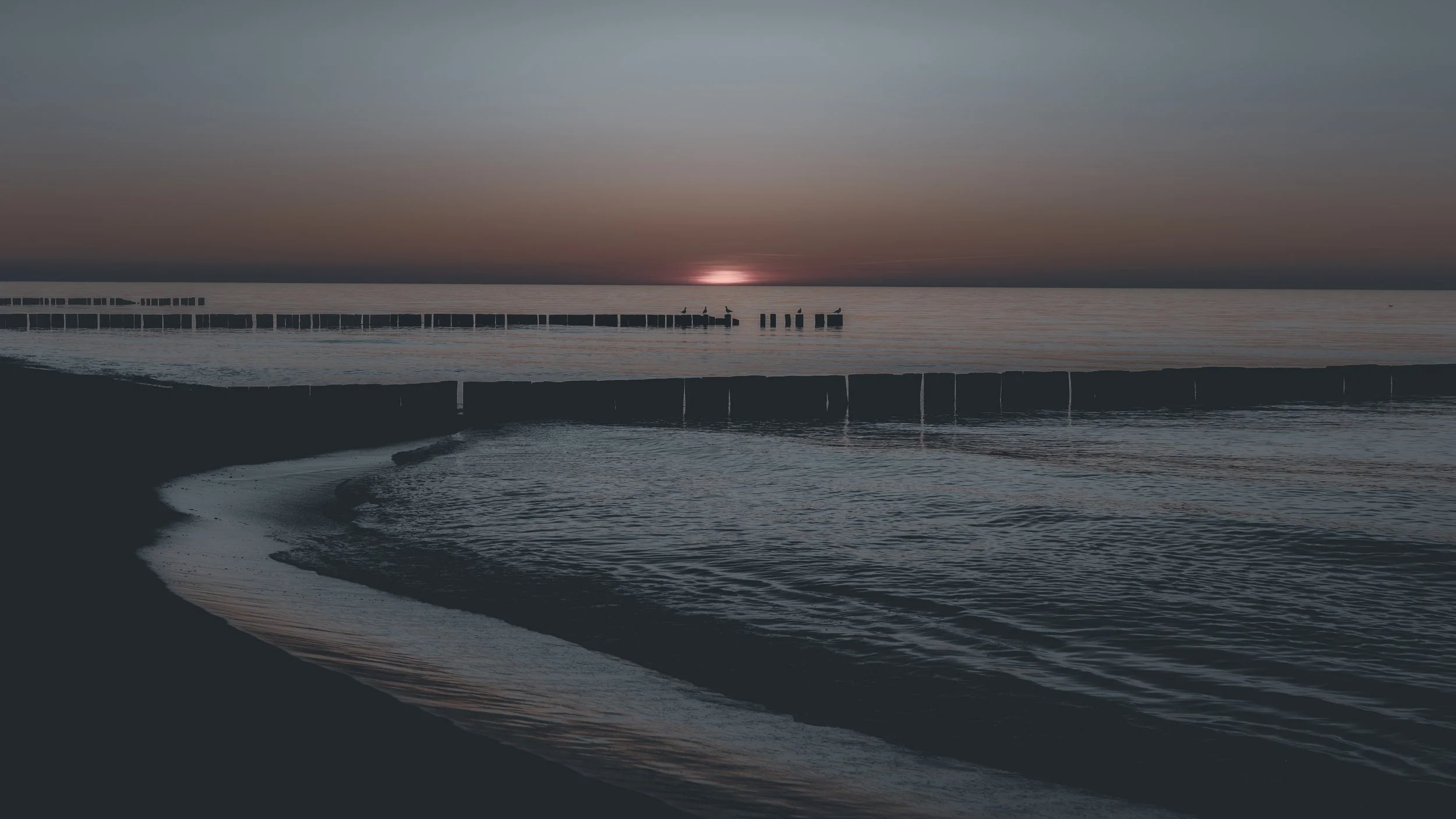 Unter dem Horizont
Nur noch ein schmaler Rest des Sonnenlichts liegt am Horizont. Buhnen, Wasser und Küstenlinie staffeln den dunkler werdenden Raum.