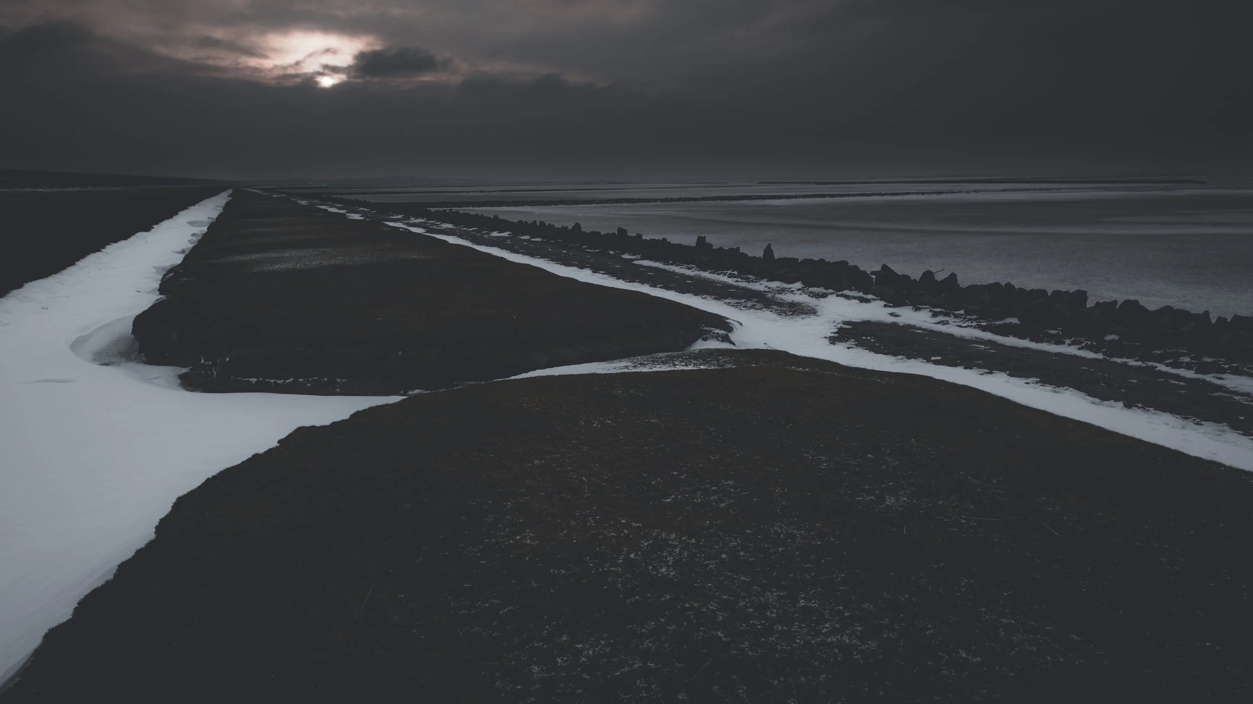 Zwischen den Linien
Schnee, Wasser und dunkles Land formen eine Küstenlandschaft aus Flächen und Linien. Das gedämpfte Licht reduziert den Raum auf seine wesentlichen Strukturen.