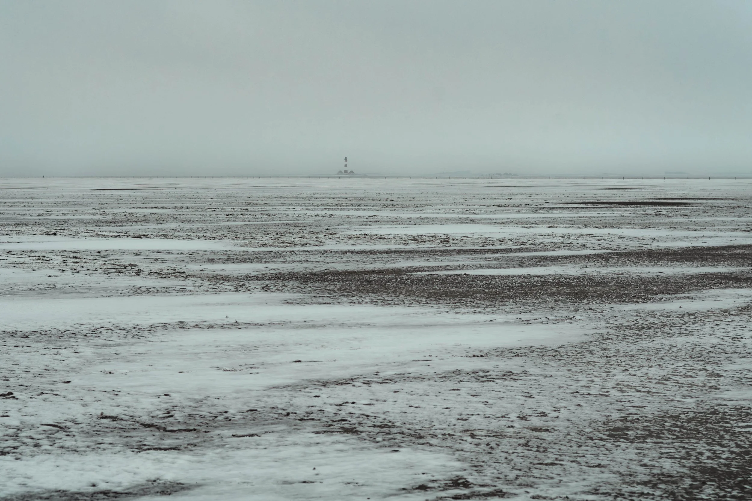 Weite winterliche Küstenlandschaft im Watt mit gefrorener Oberfläche und einem kleinen Leuchtturm am Horizont im Nebel.