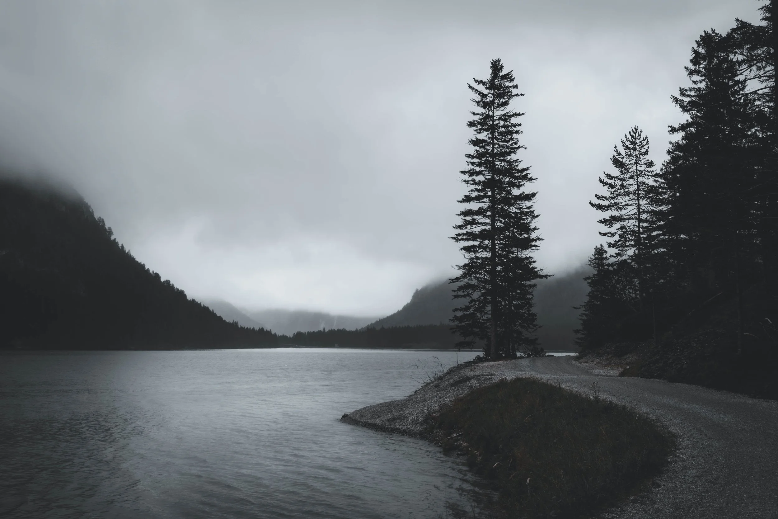 Bergsee mit Baumgruppe
Bergsee mit markanter Baumgruppe auf einer Landzunge, umgeben von bewaldeten Berghängen und tiefen Wolken – Naturfotografie von ReBlick.