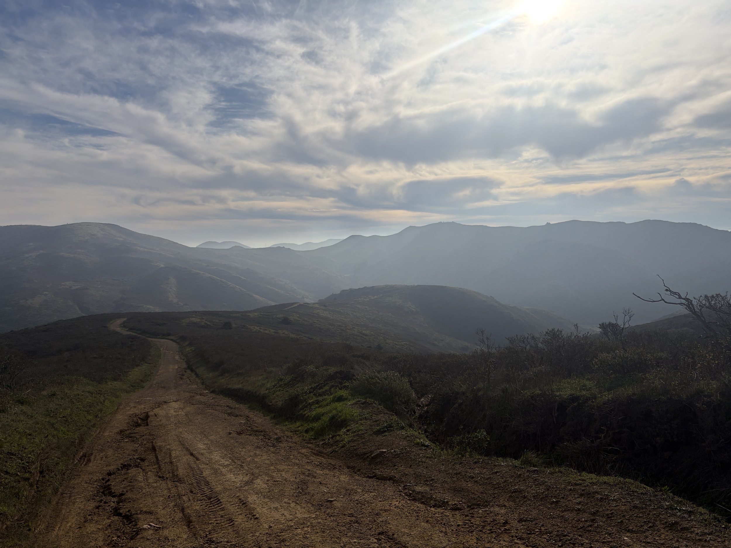Dirt trail winding through rolling hills with mountain ranges in the distance under a partly cloudy sky.