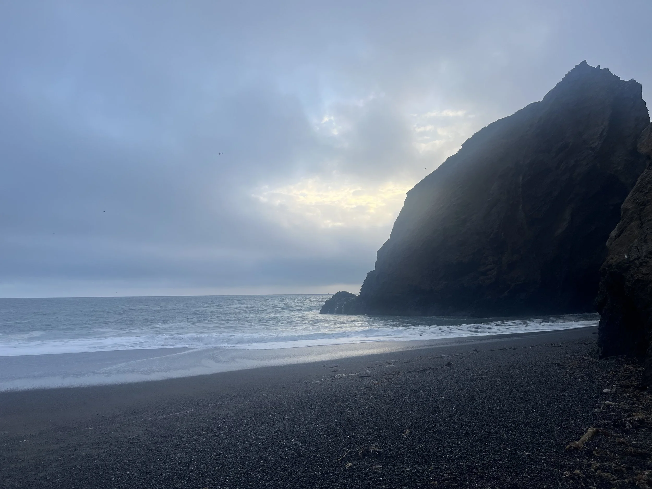 A black sand beach with a large rocky cliff on the right, cloudy sky, and a calm ocean with gentle waves