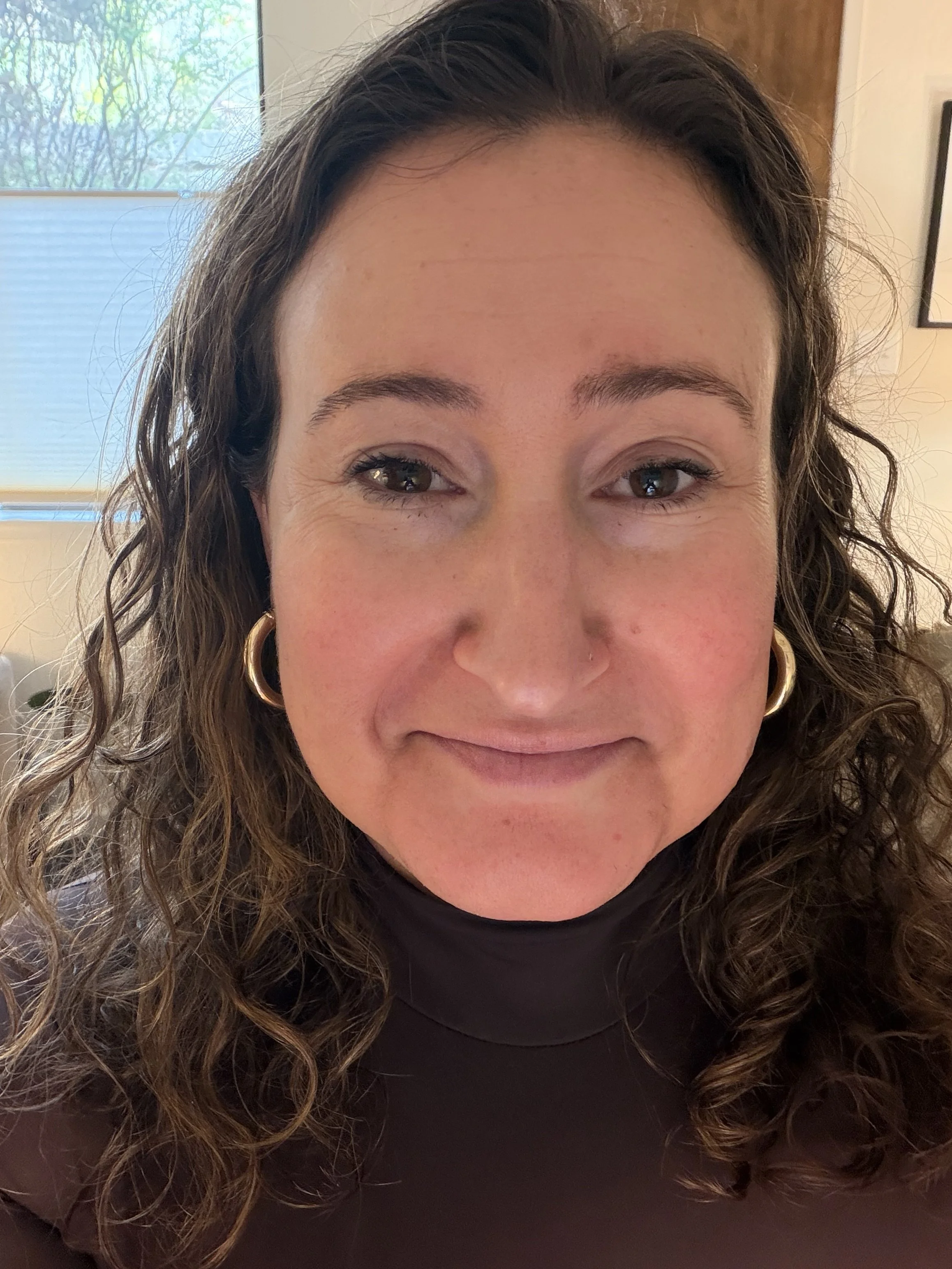 A close-up of a woman with curly brown hair, gold hoop earrings, wearing a black turtleneck, smiling softly, in a well-lit room with a window in the background.
