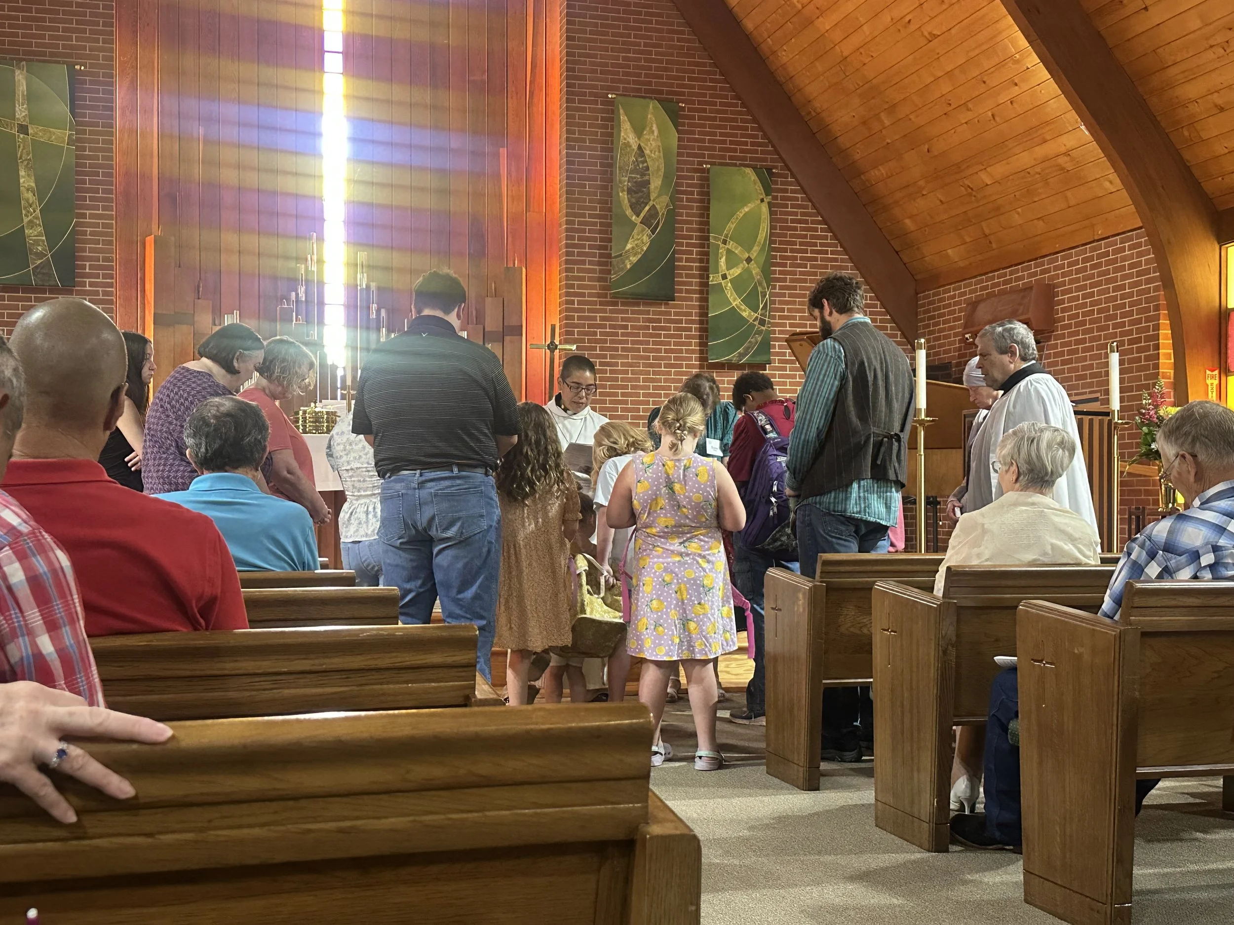 People gather in a church during a service or prayer, with some seated on wooden pews and others standing, illuminated by sunlight streaming through a tall stained glass window.