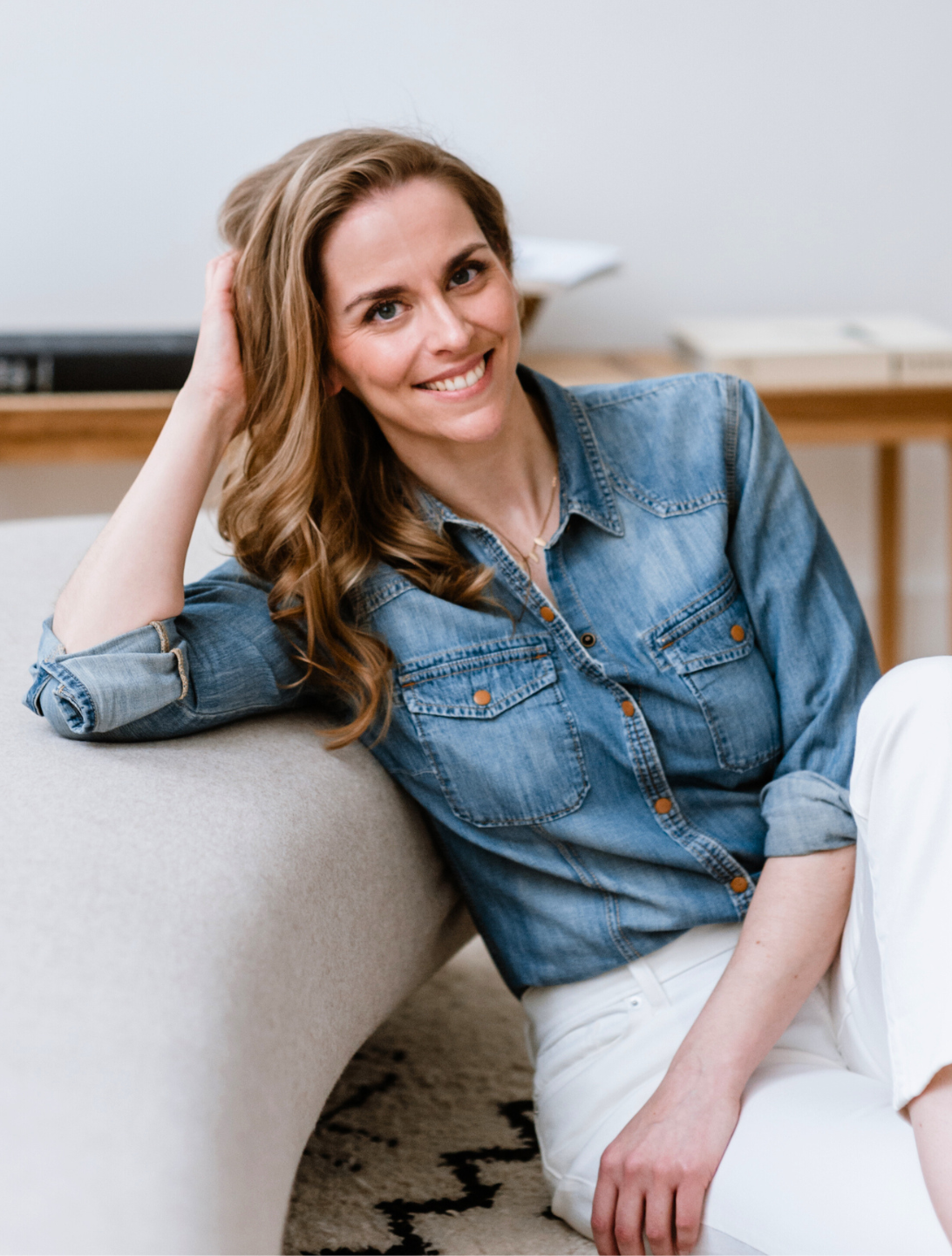A smiling woman with long, wavy brown hair sitting on a beige couch, wearing a denim shirt and white pants, in a room with a desk and books in the background.